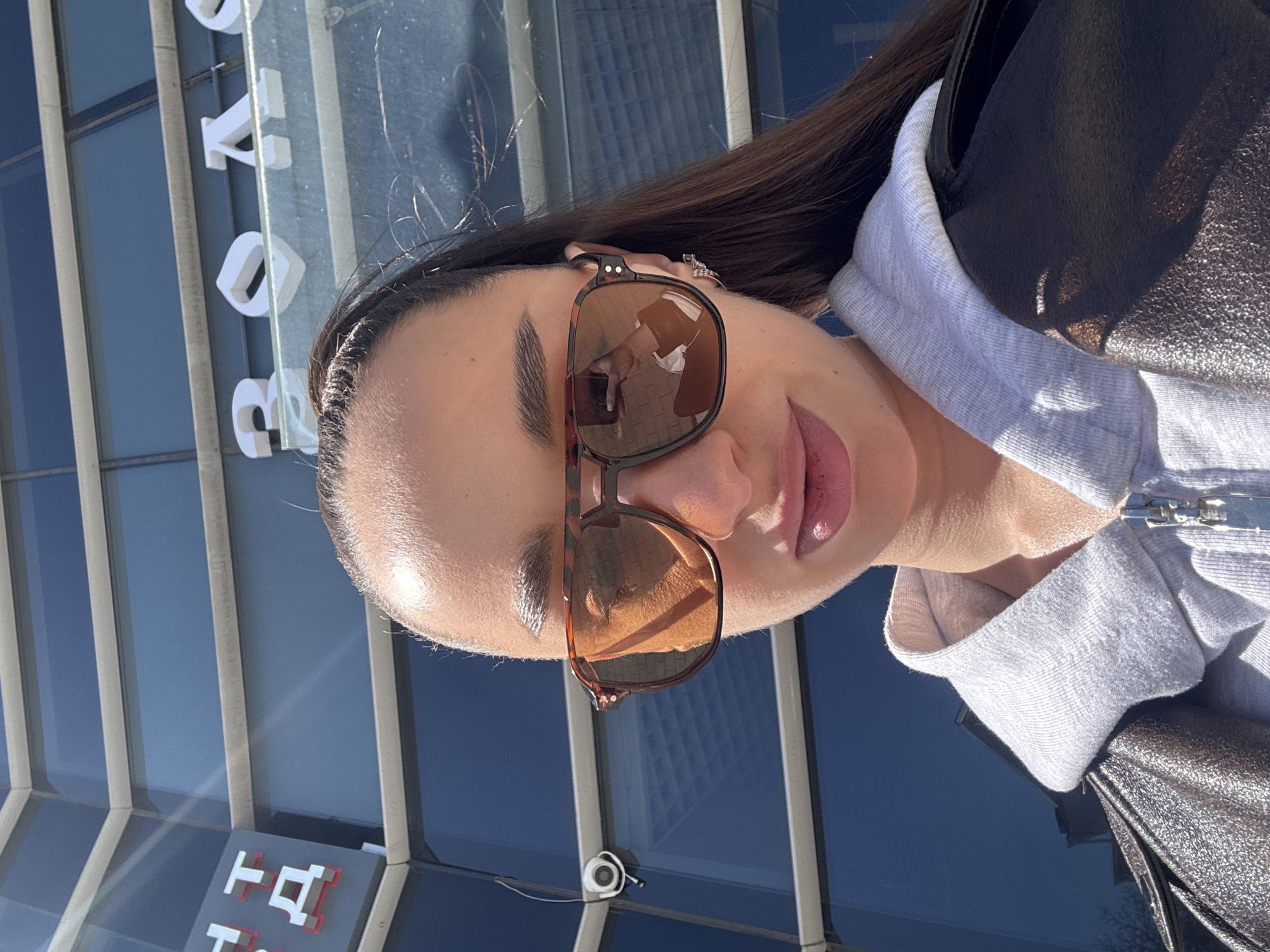 The image is a brightly lit, close-up selfie of a woman outdoors, taken during the daytime under clear, sunny weather. The woman is looking directly at the camera, wearing stylish tortoise-shell sunglasses with brown-tinted lenses. Her long dark hair is pulled back, and she has well-defined eyebrows and glossy lips. She is wearing a light grey hooded top with a zipper, under a darker, possibly metallic-textured jacket or top. Reflections in her sunglasses show a hand holding a phone, indicating the selfie was taken by herself. She also has visible earrings.
The background features a modern building with large blue-tinted windows and vertical white or light grey architectural elements. In the upper left portion of the background (when viewed upright), white three-dimensional letters spelling "ROSA" are visible on the building. Further down in the background, a small sign with white characters, including shapes resembling "T" and "Q" with red outlines, is present, alongside a small security camera. The setting suggests an urban or commercial environment in Belorechensk, Russia.