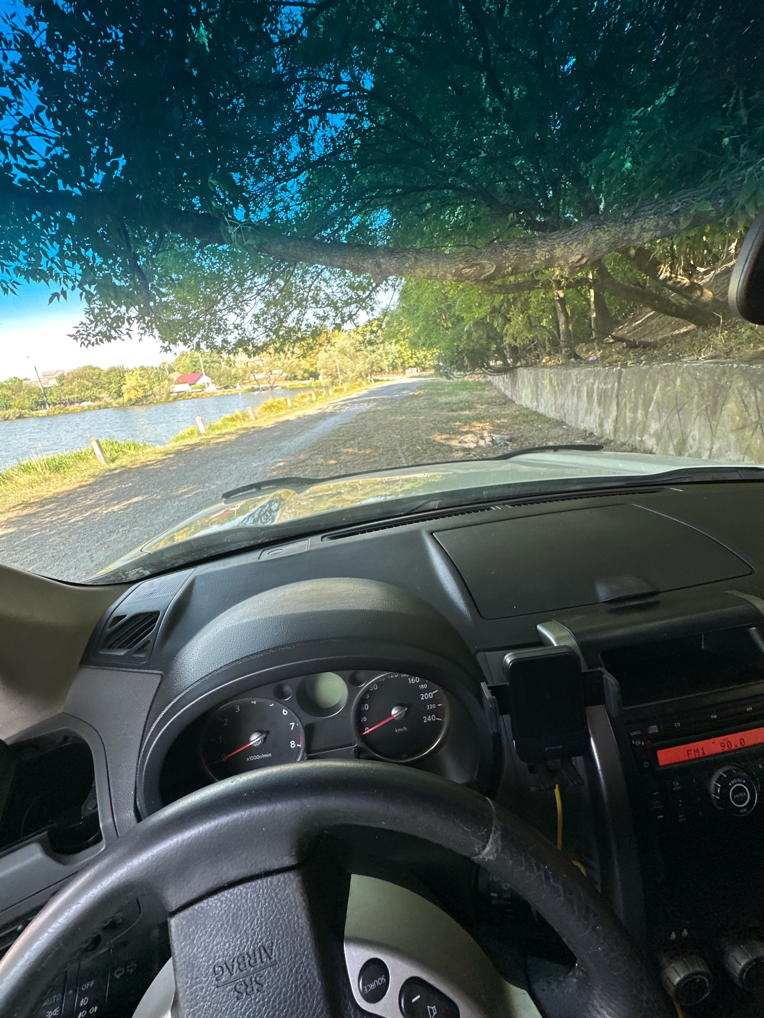 The image is taken from the interior of a car, looking out through the windshield. A large tree branch overhangs the windshield, casting dappled shadows. The scene outside the car appears to be a road next to a body of water, possibly a lake or river, with vegetation and a few buildings in the distance. The dashboard of the car is visible in the foreground, including the steering wheel, instrument cluster showing speed in km/h and RPM, and the car's radio displaying "FM1 90.0". The location is identified as Makiv, Ukraine.