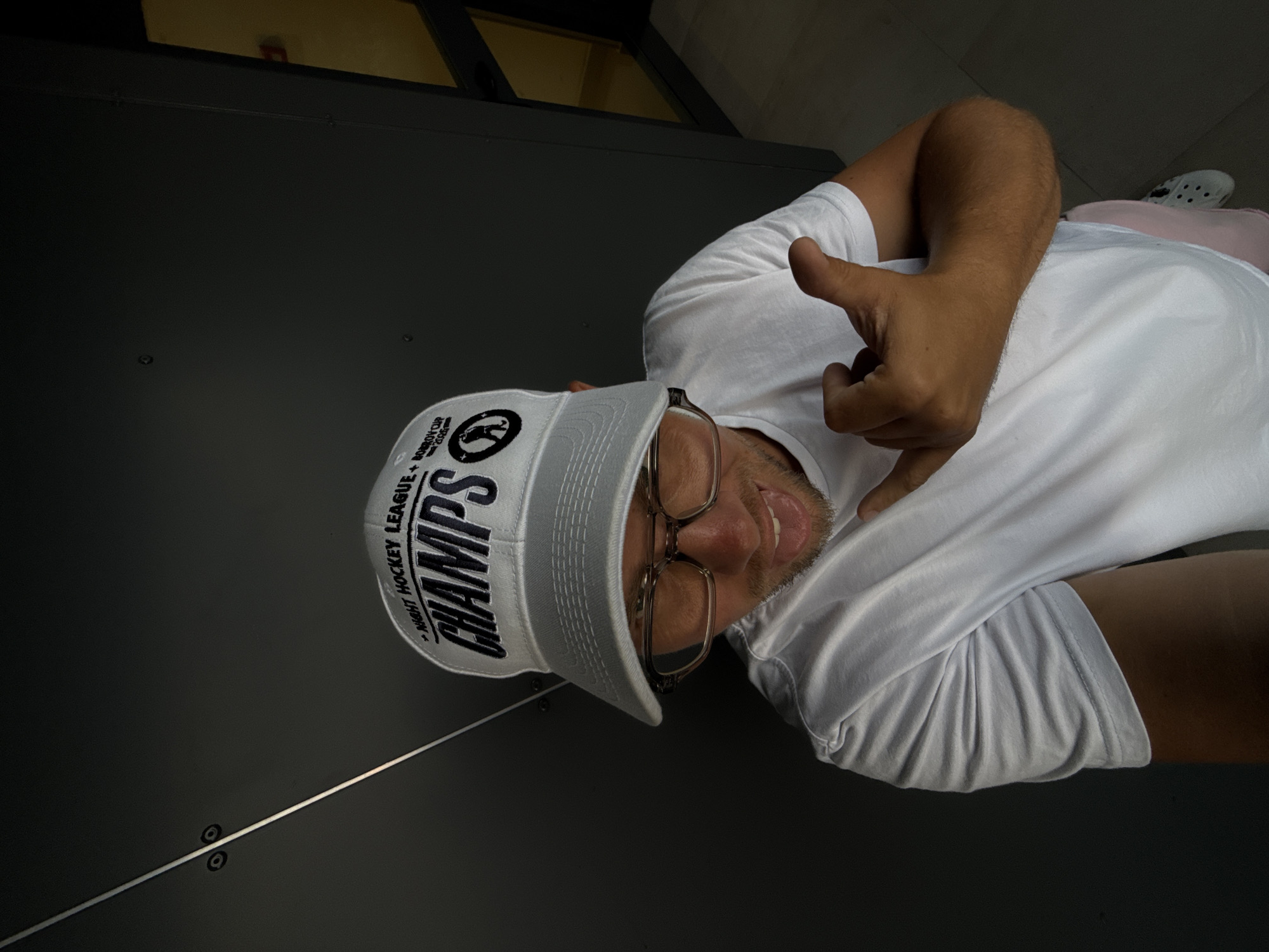 A man wearing a white t-shirt and a white baseball cap with the words "Night Hockey League Champs" and "Dobrov Cup 2025" on it is taking a selfie against a dark grey wall. He is also wearing glasses and has his tongue sticking out. The man's right arm is raised with his hand in a "shaka" sign. In the background, there is a glimpse of a yellow wall and a dark doorway. The scene appears to be an indoor setting, possibly a public space or corridor. There is no indication that this image was taken in Sochi, Russia.