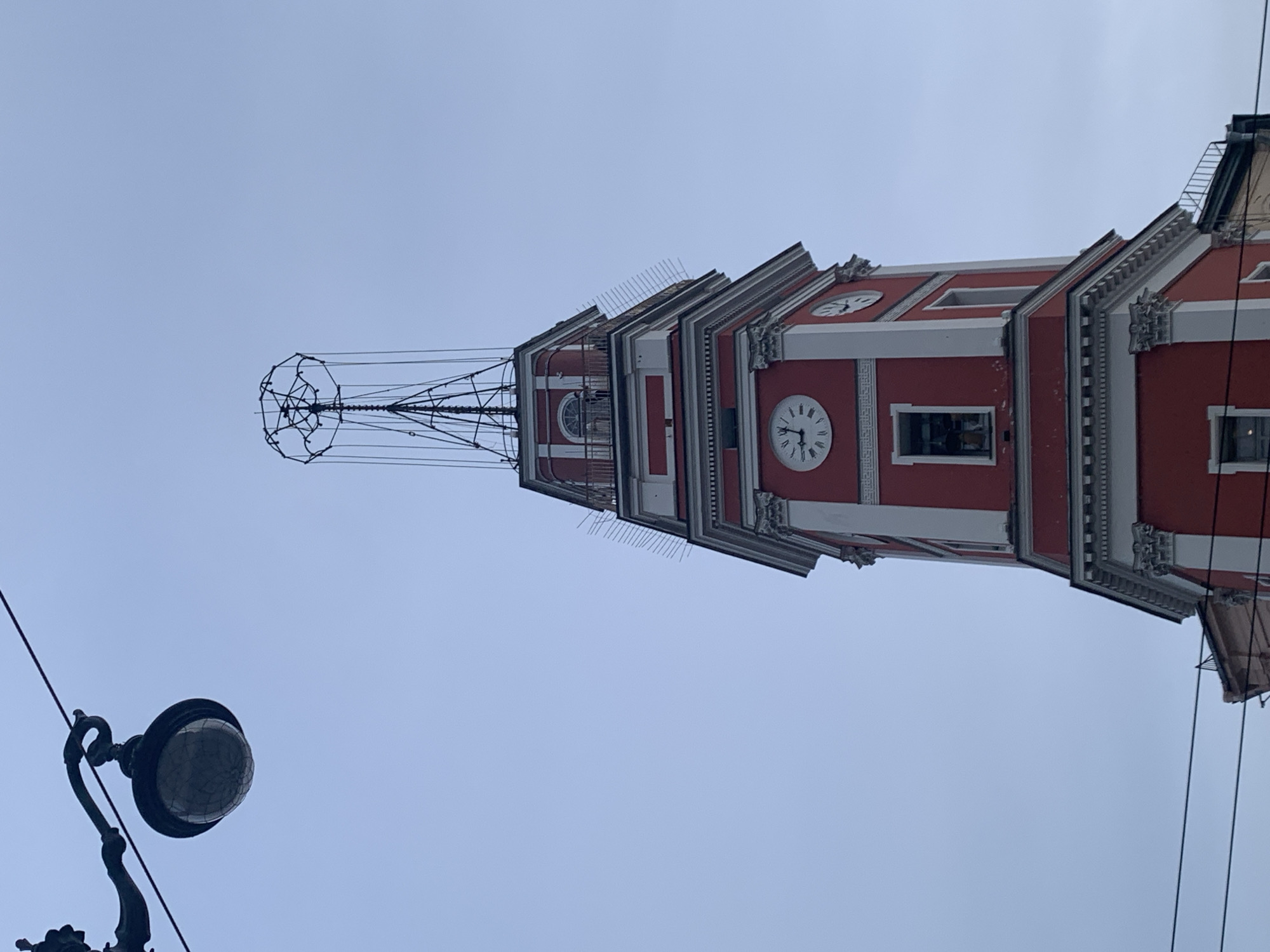 The image shows a close-up view of a red and white clock tower against a pale blue sky in Saint Petersburg, Russia. A vintage street lamp hangs in the upper left corner. The tower features classical architectural details, including columns and decorative moldings, and has multiple clock faces. The uppermost section of the tower is topped with a complex metal structure that resembles an antenna or a weather vane. A series of thin metal spikes, likely bird deterrents, are visible along the edges of some of the tower's levels. There are no people clearly visible in the frame, though a faint silhouette of a person can be seen on a balcony near the top of the tower. The image is captured from a low angle, emphasizing the height and grandeur of the structure.