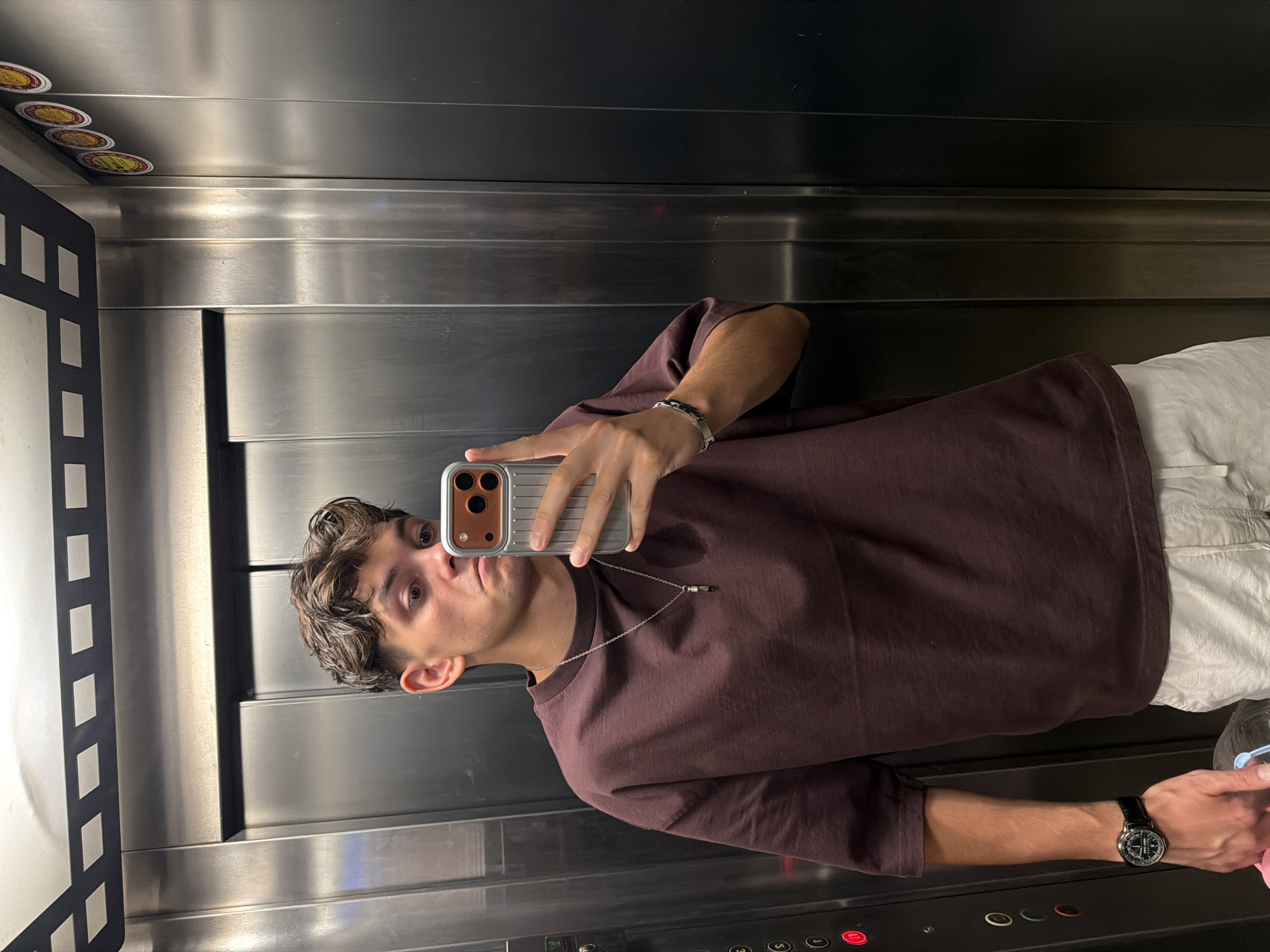 In an elevator, a young man with brown, wavy hair is taking a selfie. He is wearing a loose-fitting, dark maroon t-shirt and light-colored shorts. A silver chain with a pendant hangs around his neck, and he has a silver bracelet on his left wrist. A watch with a black strap is visible on his right wrist. He is holding a silver-cased smartphone in his right hand, angled to capture the reflection in the elevator's mirrored walls. The elevator walls are made of brushed metal, with vertical panels. There are buttons on the left side of the elevator, including a red illuminated button. The ceiling has a black decorative element with white rectangular shapes. Several round stickers are visible on the upper right wall of the elevator. The scene appears to be in Blagoevgrad, Bulgaria.