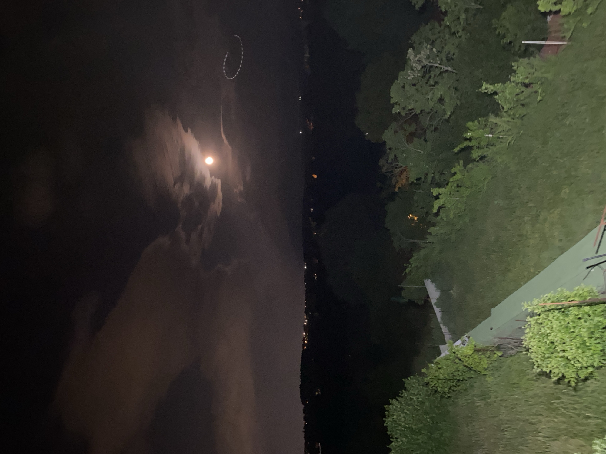 This night-time image, captured with a vertical orientation, displays a serene outdoor scene encompassing a body of water and a treelined landscape. The bright, full moon is a central subject, partially veiled by soft clouds, and its light reflects gently on the calm surface of the water on the left side of the frame.
Along the distant bank of the water body, numerous small, shimmering lights are visible, indicating the presence of a town or settlement. A notable detail in the sky, above the water, is a faint yet distinct white circular trail or light streak, adding a touch of mystery to the nocturnal vista.
The right side of the image features dark silhouettes of trees and lush green grass, suggesting a natural or cultivated landscape. In the immediate foreground, a light green or grey sloped surface, possibly a tennis court or a large roof, is partially visible, surrounded by dense bushes and other vegetation.
The weather appears cloudy, contributing to the diffuse glow around the moon. The overall impression is that of a quiet, reflective night over a lake or large river, possibly in the vicinity of Bankya, Bulgaria. No people or visible text are present in the image.