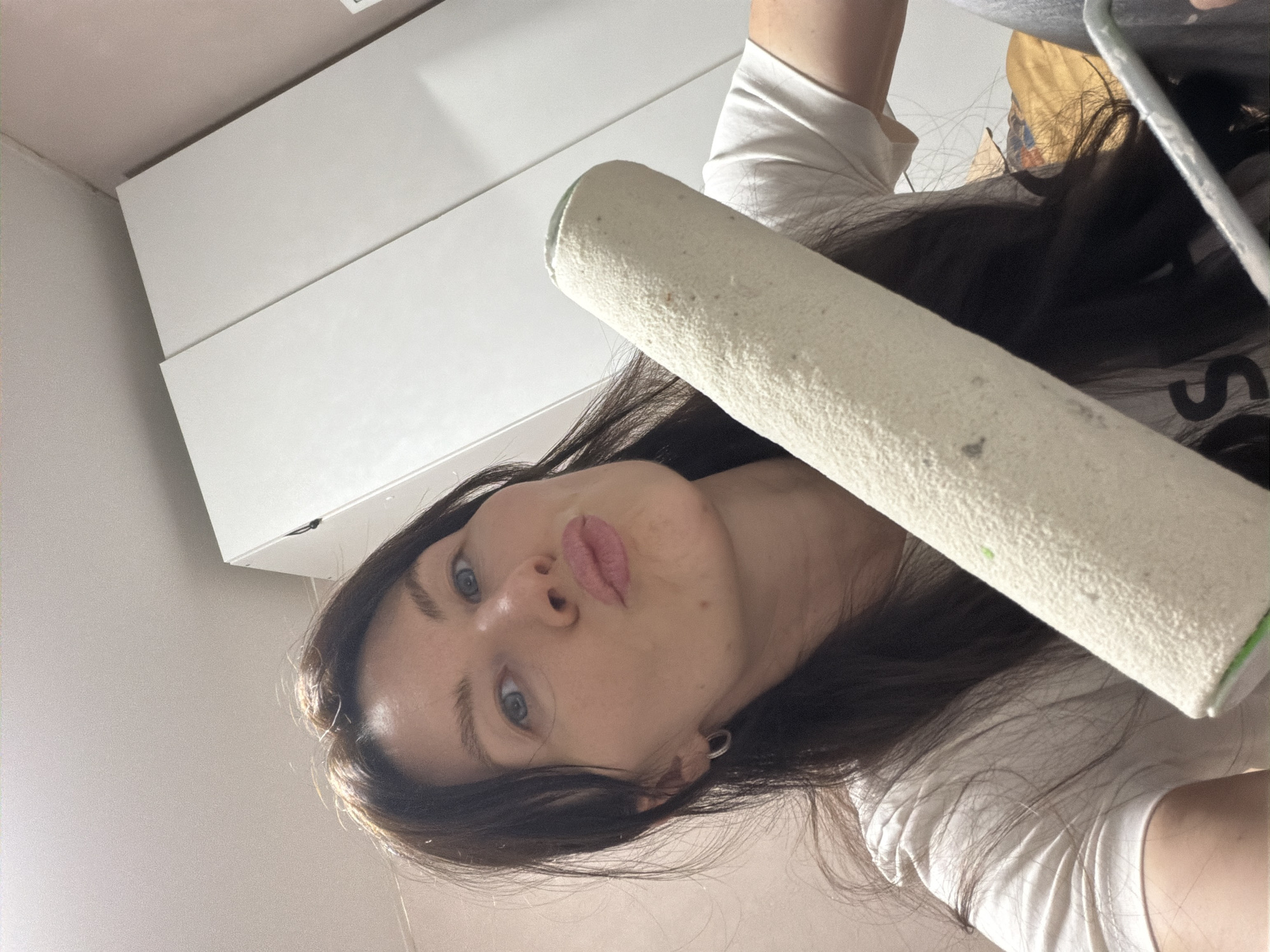 A female individual with long, dark brown hair and light blue eyes is depicted in an indoor, close-up, selfie-style photograph. She is wearing a white t-shirt and a small silver hoop earring on her visible left ear. Her lips are pursed into a "kissing" expression, and she is looking directly at the camera. A beige-colored paint roller, showing signs of use with minor specks, is held or positioned prominently in the foreground, partially obscuring her lower right side. The background consists of a white ceiling, a light pink-colored wall, and a white, two-panel wardrobe or cabinet on the right side of the frame. A blurred, patterned object is partially visible in the lower right background. The specific location within Perm, Russia, is not discernible from the provided image.