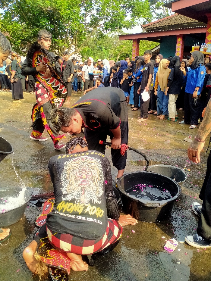 In a wet outdoor setting, a ritual unfolds with a crouching figure in a black shirt with a stylized graphic on the back, a red and white patterned garment, and wet hair. A man in black leans over, a whip in hand, near buckets of water with floating petals. Other individuals, some in traditional attire, observe, while a crowd, including some in blue jackets and headscarves, gathers in the background, suggesting a performance or ceremony is underway.