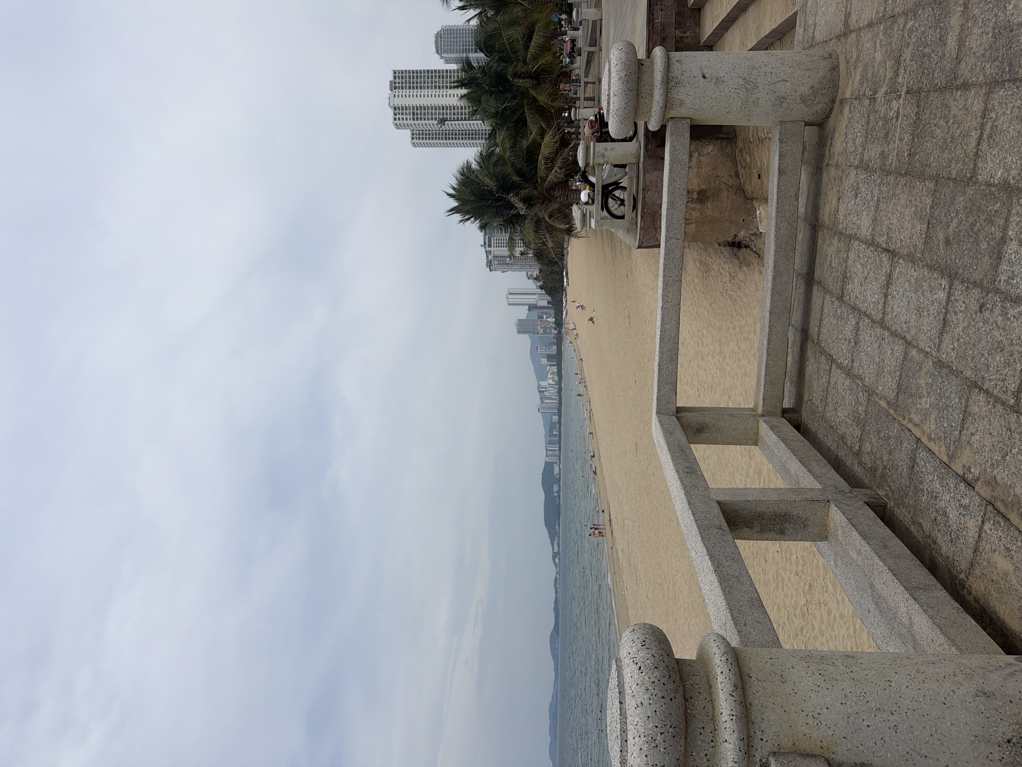 The image shows a view of Nha Trang beach in Nha Trang Ward, Vietnam, taken from an elevated walkway or promenade bordered by a low, grey stone railing. The beach itself is sandy and stretches along the coastline, with sparse figures of people visible. The sea is calm with a grey sky overhead. A line of palm trees runs parallel to the beach, separating it from the urban landscape of high-rise buildings in the background. Parked motorcycles are on the promenade near the trees. The sky is overcast, and the overall color palette is muted.