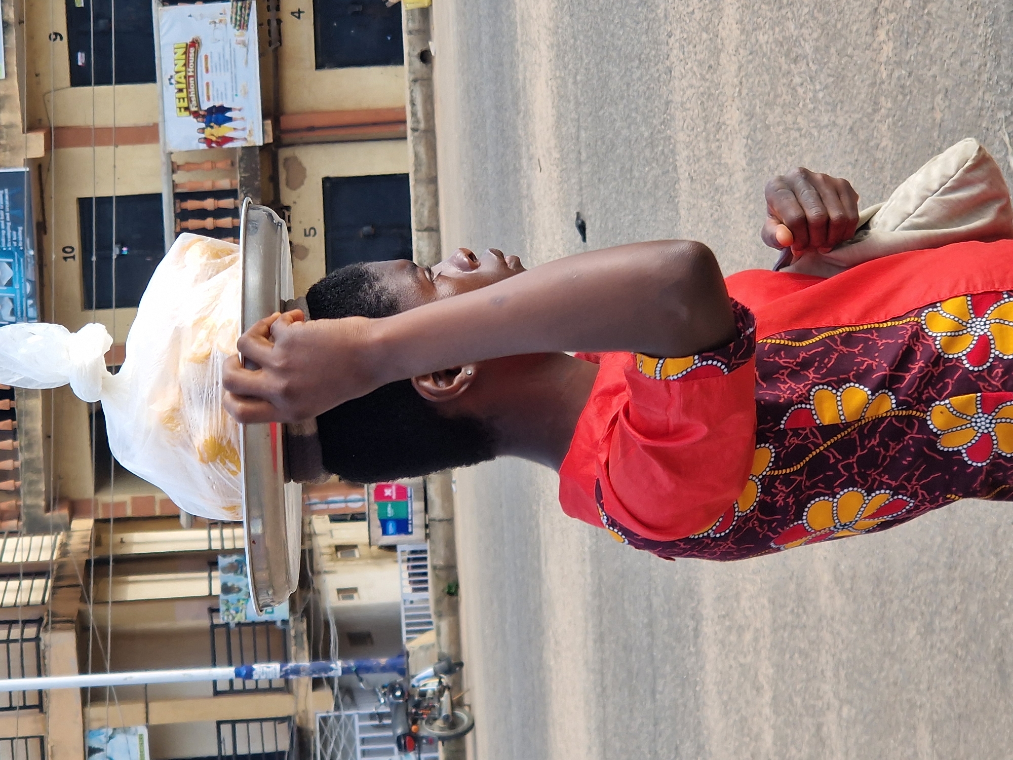 In New Stadium, Nigeria, a young woman is captured in a side profile, balancing a tray of food items wrapped in clear plastic on her head. She is wearing a vibrant red top with a patterned fabric featuring yellow and red floral designs on a dark maroon background. Her right arm is raised, stabilizing the tray, while her left hand rests on a light-colored cloth bag slung over her shoulder. The background reveals a street scene with buildings, including one with numbered units like '10' and '5', and a sign for "FELIANNI Fashion House." A motorcycle is visible on the left side of the frame, parked near a white railing. The ground is paved, indicative of a street or marketplace. The lighting suggests it is daytime.