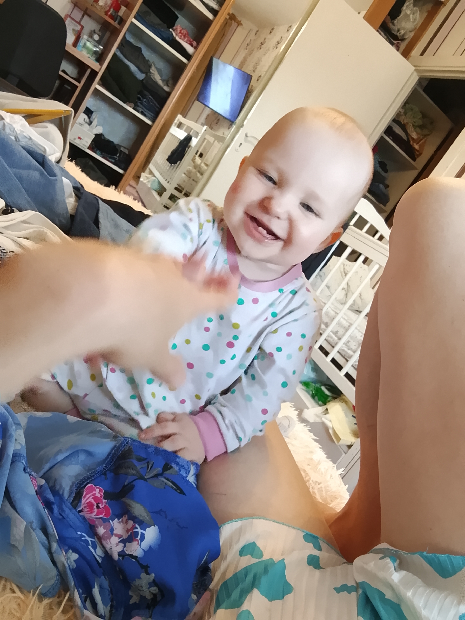 An infant is sitting on a furry rug in a room with a crib and a closet in the background. The infant is wearing a white long-sleeved shirt with colorful polka dots. The infant is smiling and looking at the camera. A person's arm and hand are visible in the foreground, reaching towards the infant. The room appears to be a bedroom or nursery, with clothing and other personal items scattered around. The closet contains shelves filled with folded clothes. A television screen is visible on a shelf in the closet. The scene suggests a casual, domestic setting. There is no indication of the location being in Minsk, Belarus.