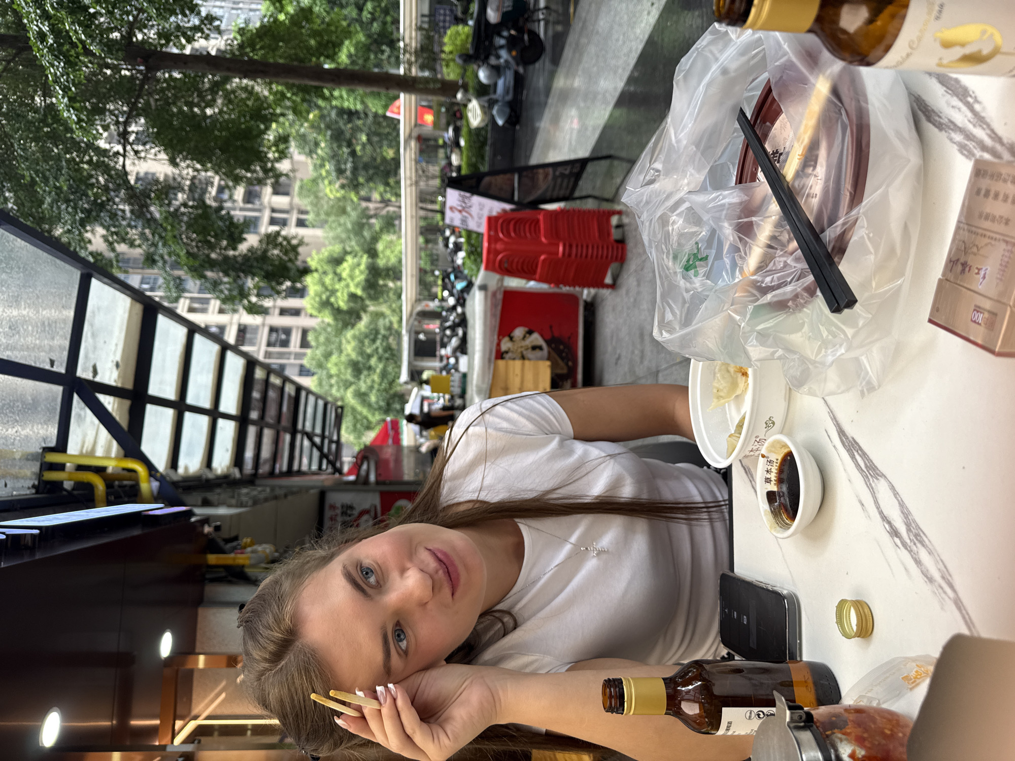 A woman with long brown hair and blue eyes sits at a table outside a restaurant. She is wearing a white t-shirt and a silver cross necklace. Her left hand rests on her forehead, and she holds chopsticks in her fingers. On the table in front of her are a bottle of beer, a bowl of dumplings, a small dish of soy sauce, a smartphone, and a pack of cigarettes. The table has a marble-like pattern. In the background, there are buildings, trees, and parked motorcycles, suggesting an urban setting in Haizhu District, China.
