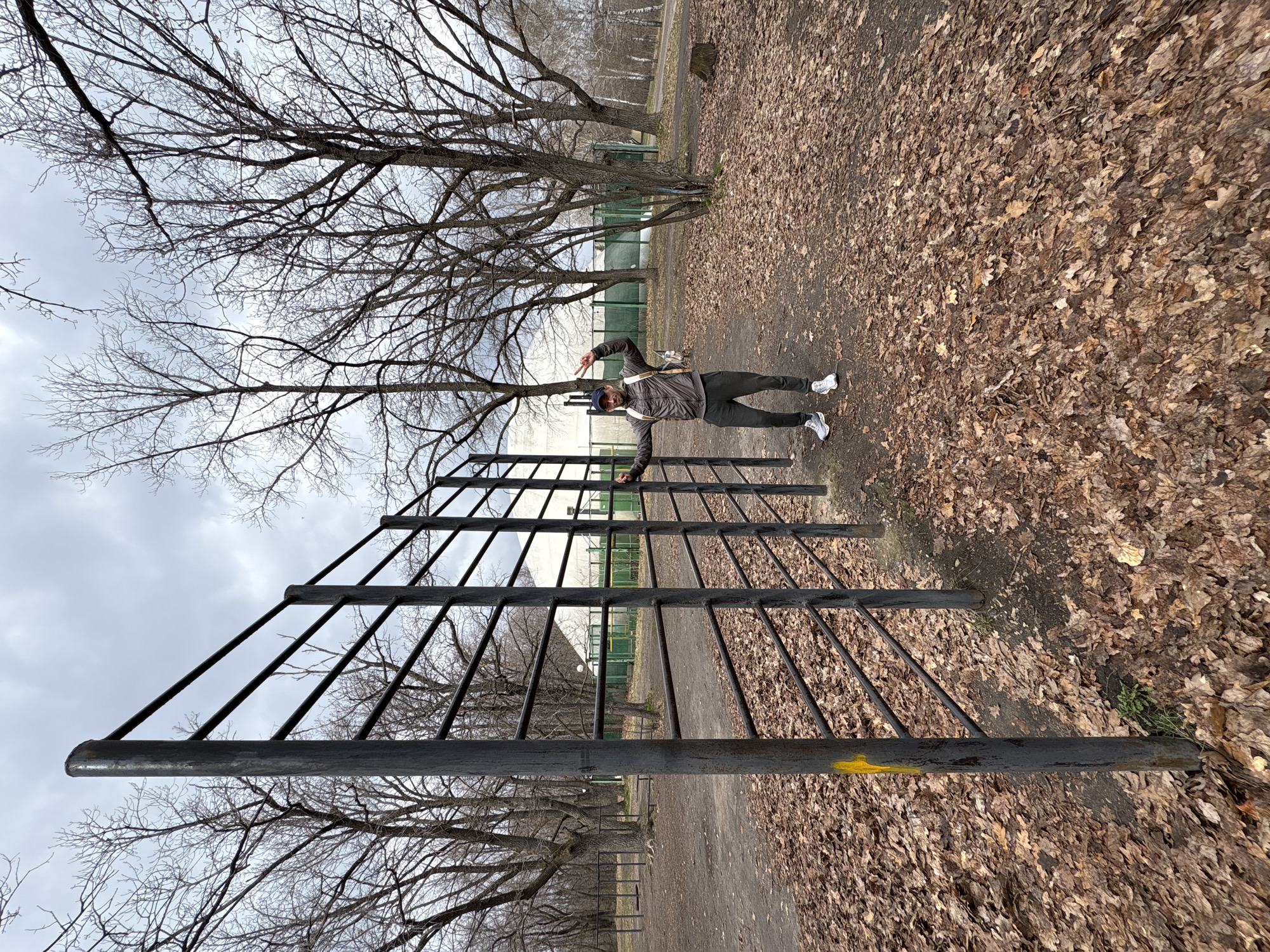 The image depicts a man standing next to a black metal ladder structure in a park-like setting. The ground is covered in fallen leaves, suggesting it is autumn or late fall. Bare trees are visible in the background, further supporting the season. The sky is overcast. The man is wearing dark pants, a jacket, and white sneakers, and a cap. He is holding his arms out and making a "V" sign with one hand. He appears to be posing for the photo. Based on the EXIF data, the location is Ulyanovsk, Russia. In the distance, there is a glimpse of a building or structure with a green fence. The ladder structure appears to be a piece of park equipment or potentially some kind of climbing frame.