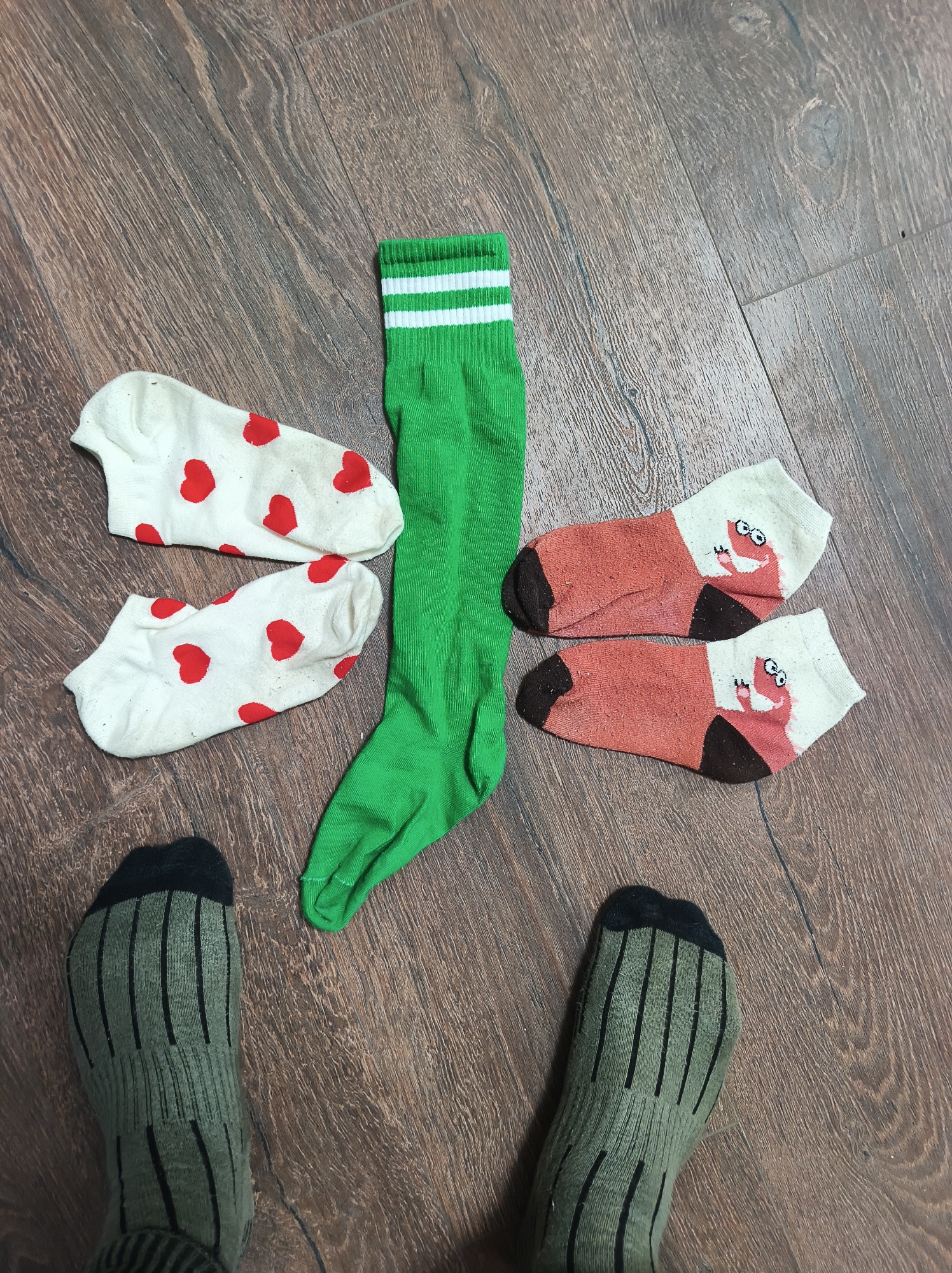 The image shows a person's feet on a wooden floor. The person is wearing olive green socks with vertical black stripes. Arranged on the floor between the person's feet are four pairs of socks: one pair is white with red hearts, another pair is green with white stripes at the top, and the third pair is white and brown with cartoon characters. The location is Vyshevychi, Ukraine.