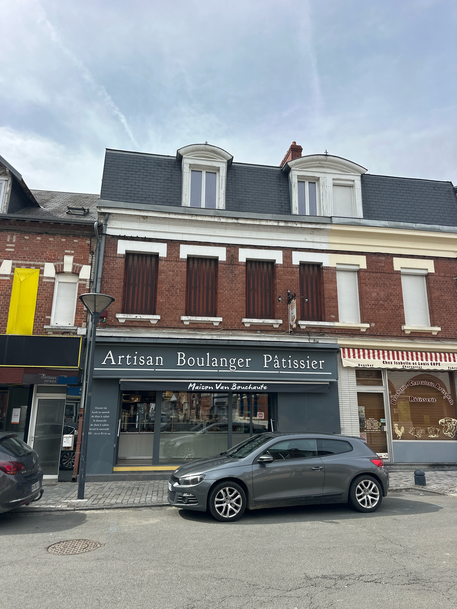 The image depicts a street scene in Roye, France, featuring the facades of two adjacent businesses. On the left, a dark grey facade displays "Artisan Boulanger Pâtissier" and "Maison Van Bouchaute" in white lettering. Below this, a glass door reveals a glimpse of the interior. To the right of the bakery, a store with a striped awning displays signage including "Chez Isabelle et Louis COPE," "Boucher," "Charcuterie Artisanale," and "Rotisserie." A gray Volkswagen Scirocco is parked in front of the bakery. The buildings have brick exteriors and dark gray mansard roofs with dormer windows. The sky is overcast, and the street is paved with cobblestones in front of the businesses and asphalt further out.