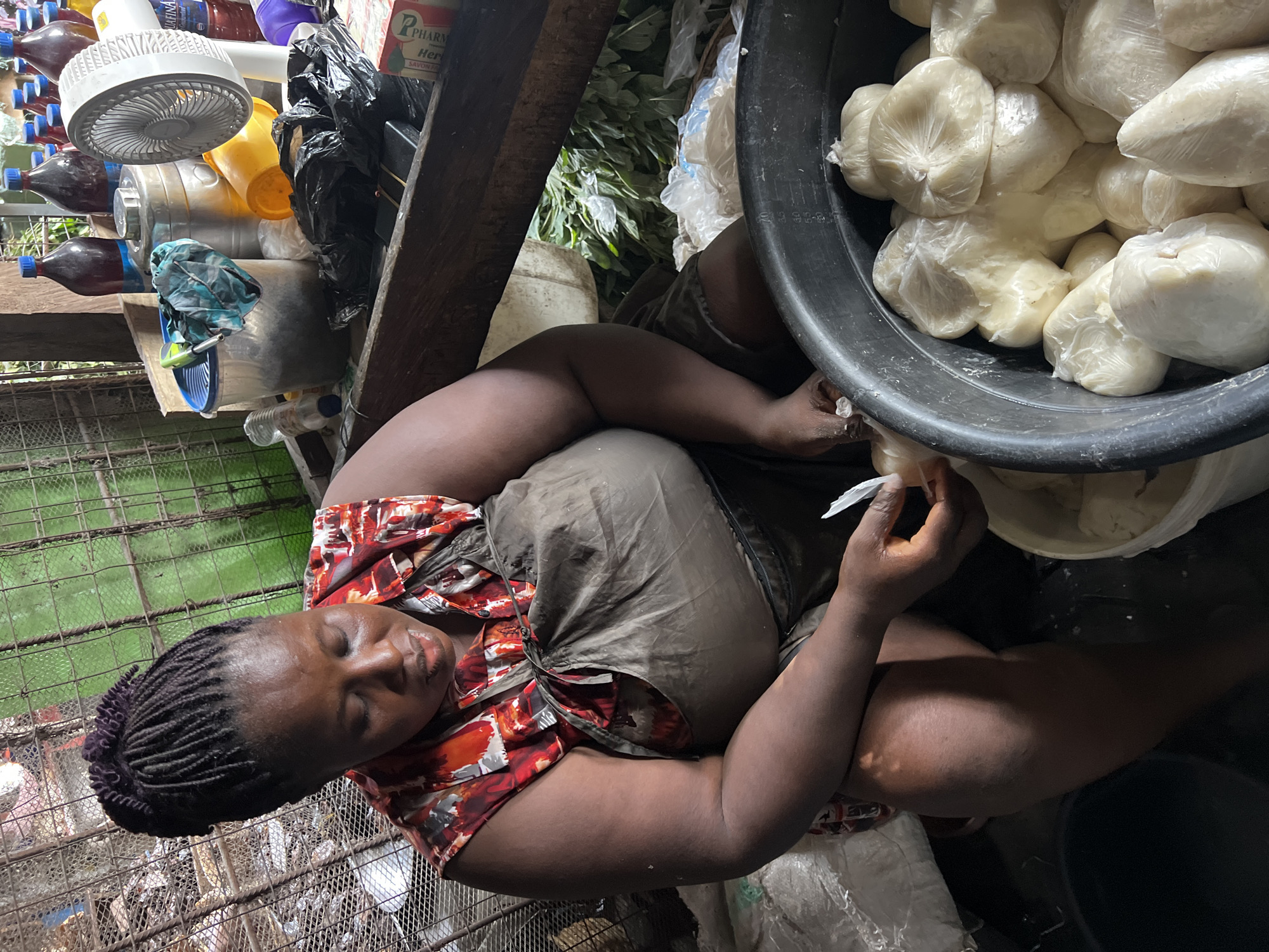 The image, taken in Nkpor, Nigeria, depicts a woman sitting on a low surface, likely the ground, within what appears to be an indoor market stall or small shop. She is a Black woman with dark braided hair, wearing a colorful, patterned short-sleeved top and a dark wrapper or skirt. Her eyes are closed, suggesting she is resting or sleeping.
To her right, a large black plastic basin is filled with numerous individually packaged portions of a white, pasty food substance, likely fufu or pounded yam, each wrapped in clear plastic. Her right hand is near or holding one of these packages, while her left arm rests on her lap.
The surroundings are densely packed with various market goods. Behind and to her left, a wooden shelf holds an assortment of items, including plastic bottles, some containing dark liquids, and a white portable electric fan. A black plastic bag and other containers are also visible. Above the fan, a reddish-brown box is partially visible, displaying the text "P PHARM HERB SAVON EX," which appears to be a label for a soap or herbal product. Further back, leafy green foliage can be seen through a gap in the structure. The ground beneath the woman appears to be a metal grid or grate. The overall lighting is somewhat dim, suggesting an indoor setting with possibly limited natural light.