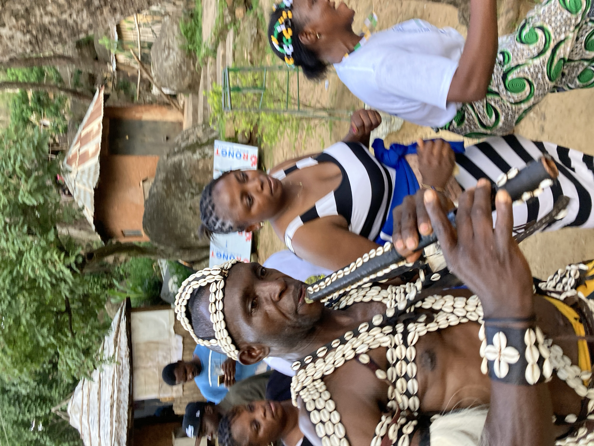 In the city of Jos, Nigeria, a group of people are gathered in an outdoor setting that appears to be a village or cultural site, with traditional huts visible in the background. A man in the foreground, adorned with numerous cowrie shells on his head, chest, and arms, plays a dark-colored wind instrument, possibly a flute or a horn. He is wearing a black wristband decorated with cowrie shells in a floral pattern. Beside him, to the right, a woman in a white t-shirt and a patterned skirt with green swirls is partially visible, her mouth open as if she is speaking or singing. To the left of the man, a woman with braided hair wears a black and white striped tank top, her arm bent at the elbow. Further in the background, other individuals are present, including a man in a blue t-shirt looking at his phone and another person wearing a baseball cap. The environment is characterized by rocky terrain, lush green trees, and rustic structures, suggesting a rural or semi-rural location within Jos. The overall scene captures a moment of cultural expression or celebration.