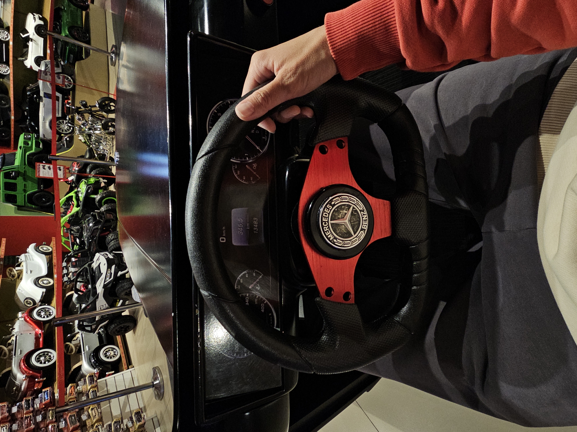 The image shows a close-up of a person's hand, wearing a red sweater, holding a black steering wheel with a Mercedes-Benz logo and red accents. The steering wheel is part of a racing simulator game, as indicated by the dashboard with gauges and a digital display. The scene is set indoors, likely in an arcade or entertainment center, given the presence of multiple toy cars displayed vertically in the background. The toy cars are of various types, including a green truck, white and red cars, and what appear to be off-road vehicles.
The digital display on the dashboard shows "0 km/h" and "345.5 km 13483". The gauges appear to represent speed and possibly RPM. The overall impression is one of engagement with a video game, suggesting a recreational activity. The lighting is artificial and suggests it could be daytime or evening. There are no clear indicators of weather or specific emotions, beyond the focused grip on the steering wheel. The location context of Moscow, Russia, is provided, but no specific landmarks or cues within the image confirm this. The vertical display of toy cars creates a unique and colorful backdrop to the central activity of playing the racing game.