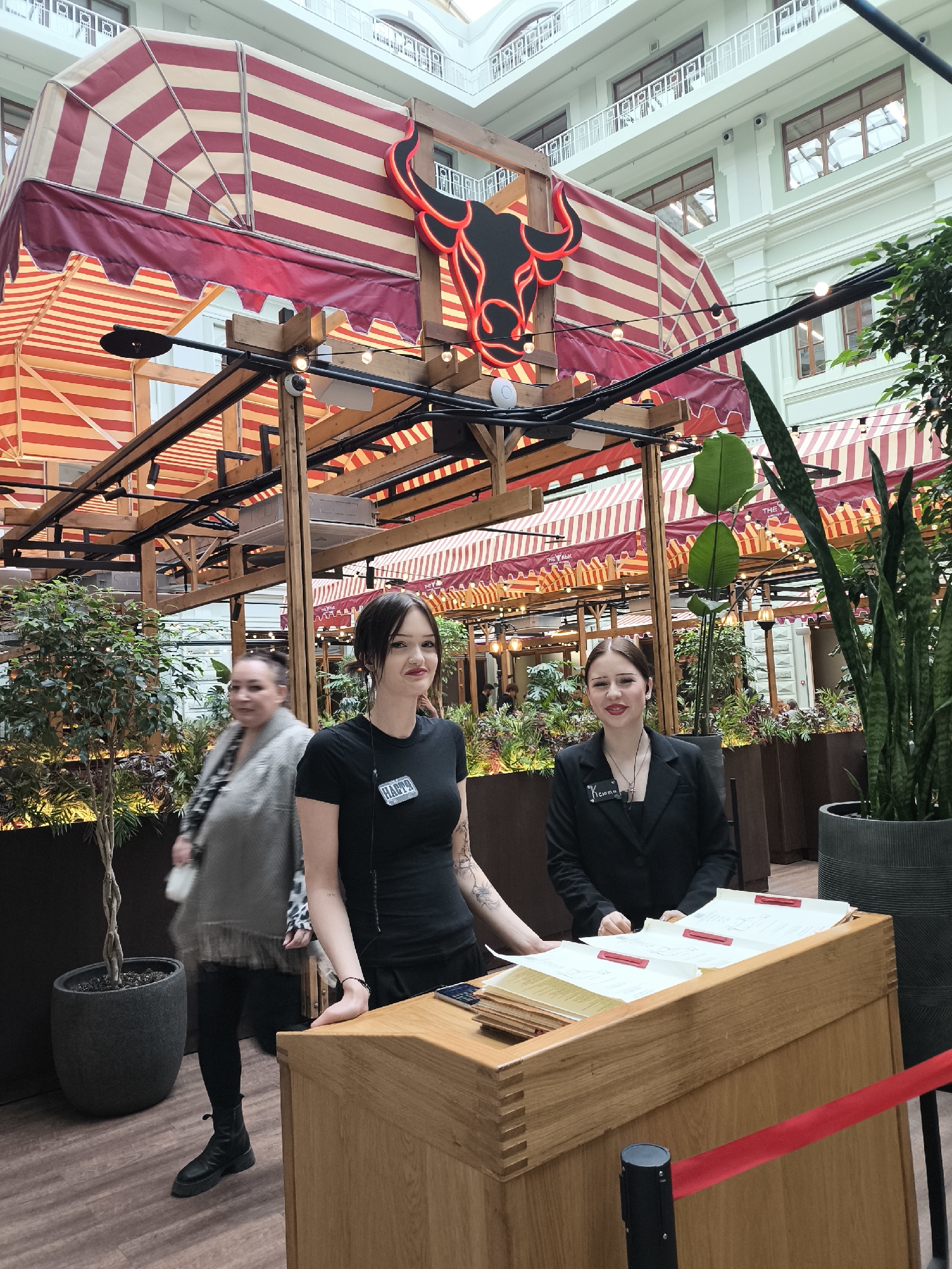 The image depicts an indoor setting, possibly a restaurant or event venue, characterized by a blend of industrial and natural elements. Two young women stand behind a wooden reception desk, likely acting as greeters or hostesses. The woman on the left wears a black t-shirt with a name tag and has visible tattoos on her arm. The woman on the right wears a black blazer, also with a name tag. The desk is adorned with papers held by red clips, suggesting reservation lists or guest information.
Behind the women is a partial view of an architectural structure featuring red and white striped awnings, reminiscent of a market stall or outdoor cafe setting. A neon sign in the shape of a bull's head is prominently displayed above the awnings. The space is decorated with greenery, including potted trees and plants, which adds a touch of nature to the indoor environment.
In the background, a woman walks towards the left side of the frame, partially obscured. The building's interior features high ceilings and architectural details, indicating a potentially historic or renovated space. Electrical cords and lights are visible overhead, suggesting ongoing activity or event preparation. The location's exact city is not known as the city is listed as Unknown, Unknown.
