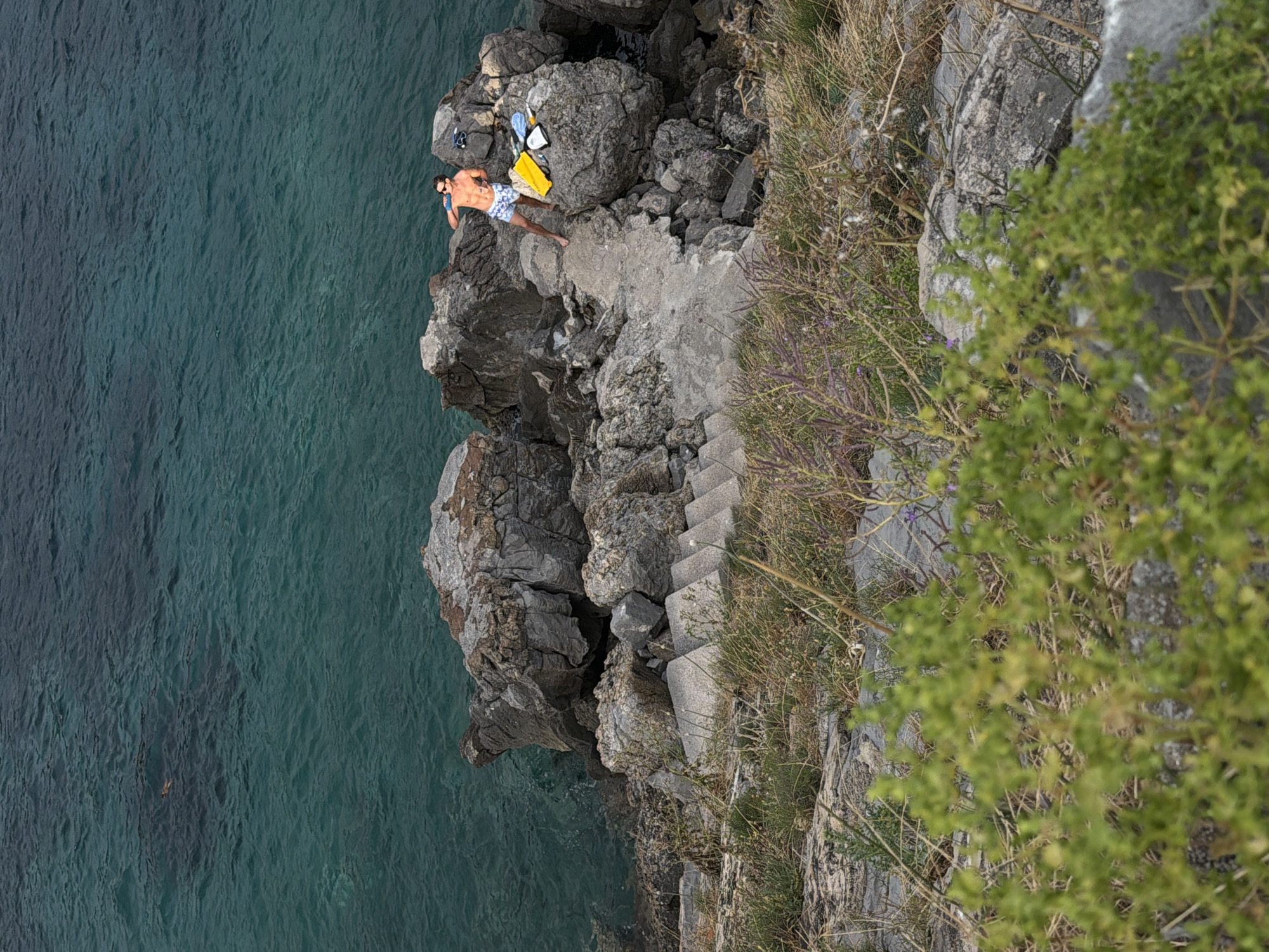 The media file displays a man in swimwear lying on large rocks next to the sea. He is positioned on a concrete staircase leading down to the water. A yellow bag and other items are placed beside him on the rocks. The sea is a deep blue-green, with some ripples on its surface. The surrounding area is rocky and overgrown with dry grass and some sparse green vegetation. The scene appears to be a natural setting, likely a coastline, with a man relaxing near the water. The lighting suggests it could be daytime, possibly late afternoon or early morning given the soft shadows. The overall mood is one of tranquility and leisure.