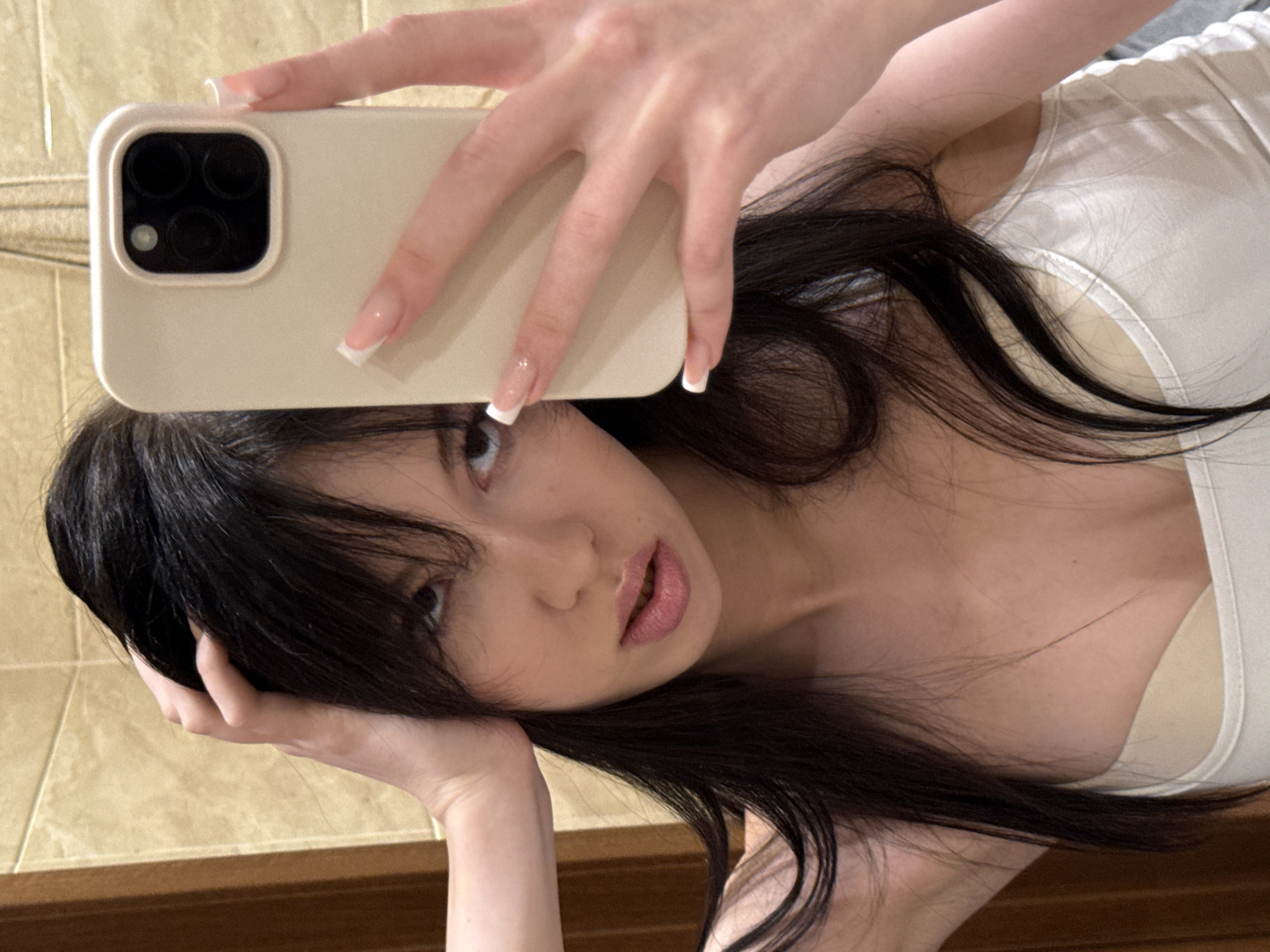 In a bathroom with beige tiled walls, a young woman with long, dark hair, taking a selfie with her phone, is partially visible. She is wearing a white tank top and a beige bra. Her right hand is holding the phone, and her left hand is resting on her head. Her facial expression is sultry, with her lips slightly parted and her eyes looking towards the camera. Her nails are manicured with white tips. The background features a wooden door and part of a mirror.