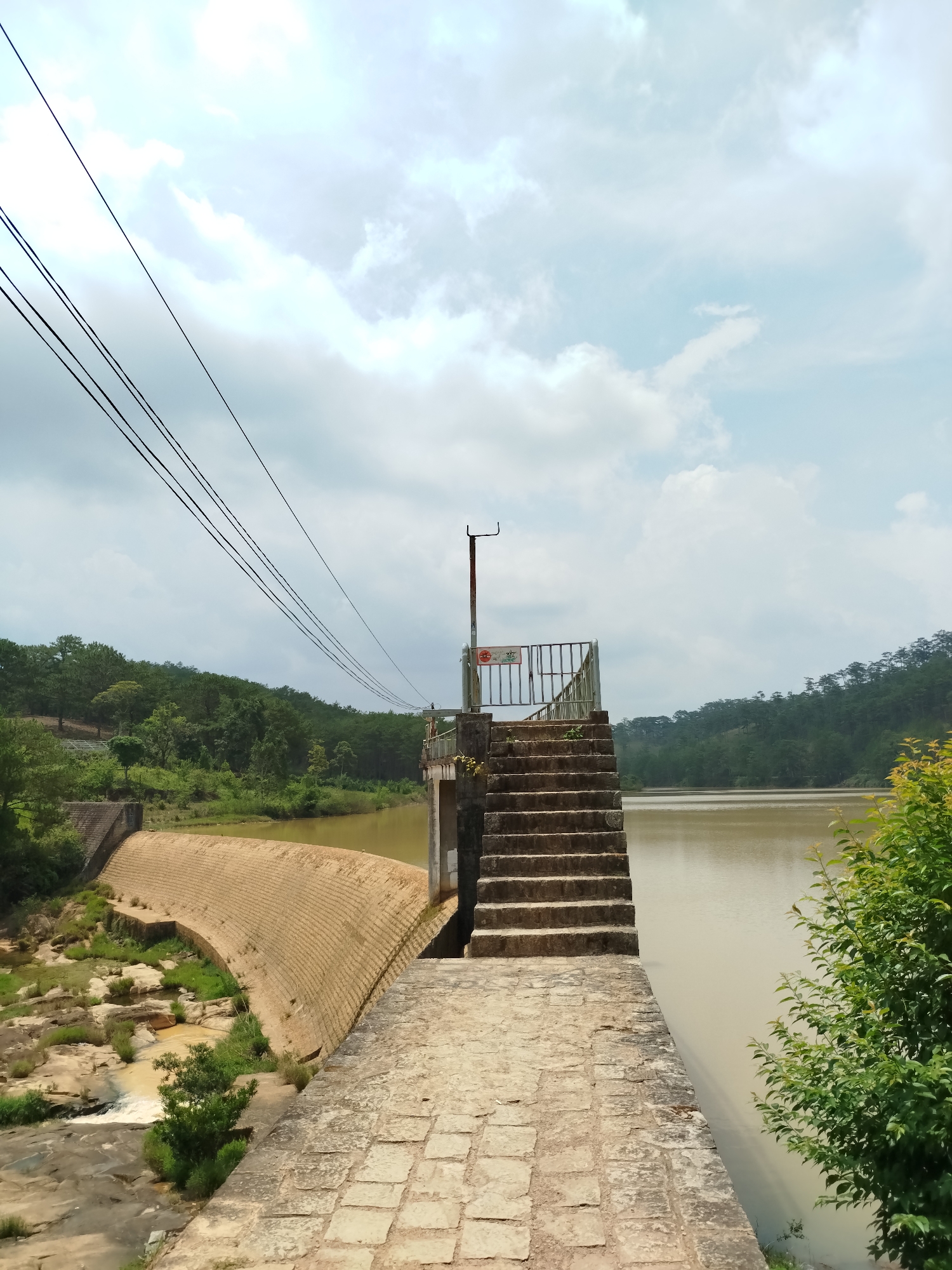 The image presents a panoramic view of a dam and reservoir in Lang Biang - Da Lat, Vietnam, under a bright, cloudy sky.
In the foreground, a stone-paved walkway leads to a set of concrete steps that ascend to a small observation platform. This platform is equipped with a metal railing and a tall, slender metal pole. To the left, a large, curved concrete dam wall holds back a body of murky, brown-toned water. Below the dam, the riverbed is visible with rocks, patches of green vegetation, and a small stream of water flowing.
The reservoir stretches across the right side of the frame, its surface reflecting the overcast sky. On the opposite bank and extending into the background, dense green forests cover rolling hills, suggesting a natural and expansive landscape. Several dark power lines traverse the upper left quadrant of the image, appearing to connect to a point near the observation platform.
Visible details include a red circular sign on the metal pole on the platform, featuring a "no entry" or "no activity" symbol over what appears to be a figure. A rectangular sign on the railing below it displays text, likely a warning, that reads "KHÔNG NHẢY" (Do Not Jump) and shows a symbol of a person diving with a red prohibition circle. The time of day is daytime, with diffused light from the cloudy sky. The scene is devoid of people, contributing to a serene and somewhat isolated atmosphere.