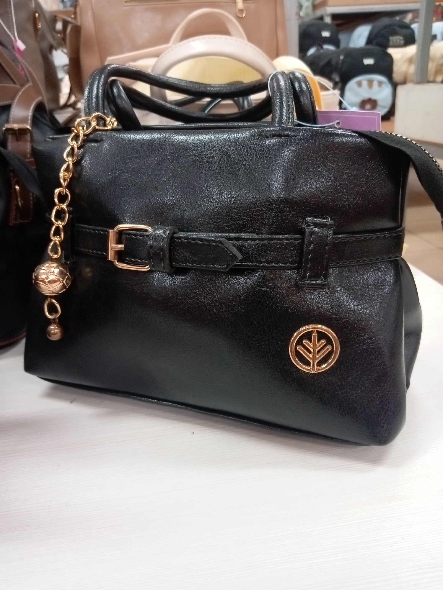 A black leather handbag with gold hardware is displayed on a white wooden surface. The handbag has a structured shape with two top handles and a decorative strap across the front with a buckle closure. A gold chain with a spherical charm and a smaller ball pendant hangs from the left handle. A round gold emblem with a stylized tree design is affixed to the front of the bag. In the background, other bags and items are visible, suggesting a retail environment. The scene appears to be an indoor display, possibly in a shop in Batam, Indonesia, though no definitive Batam-specific markers are present.