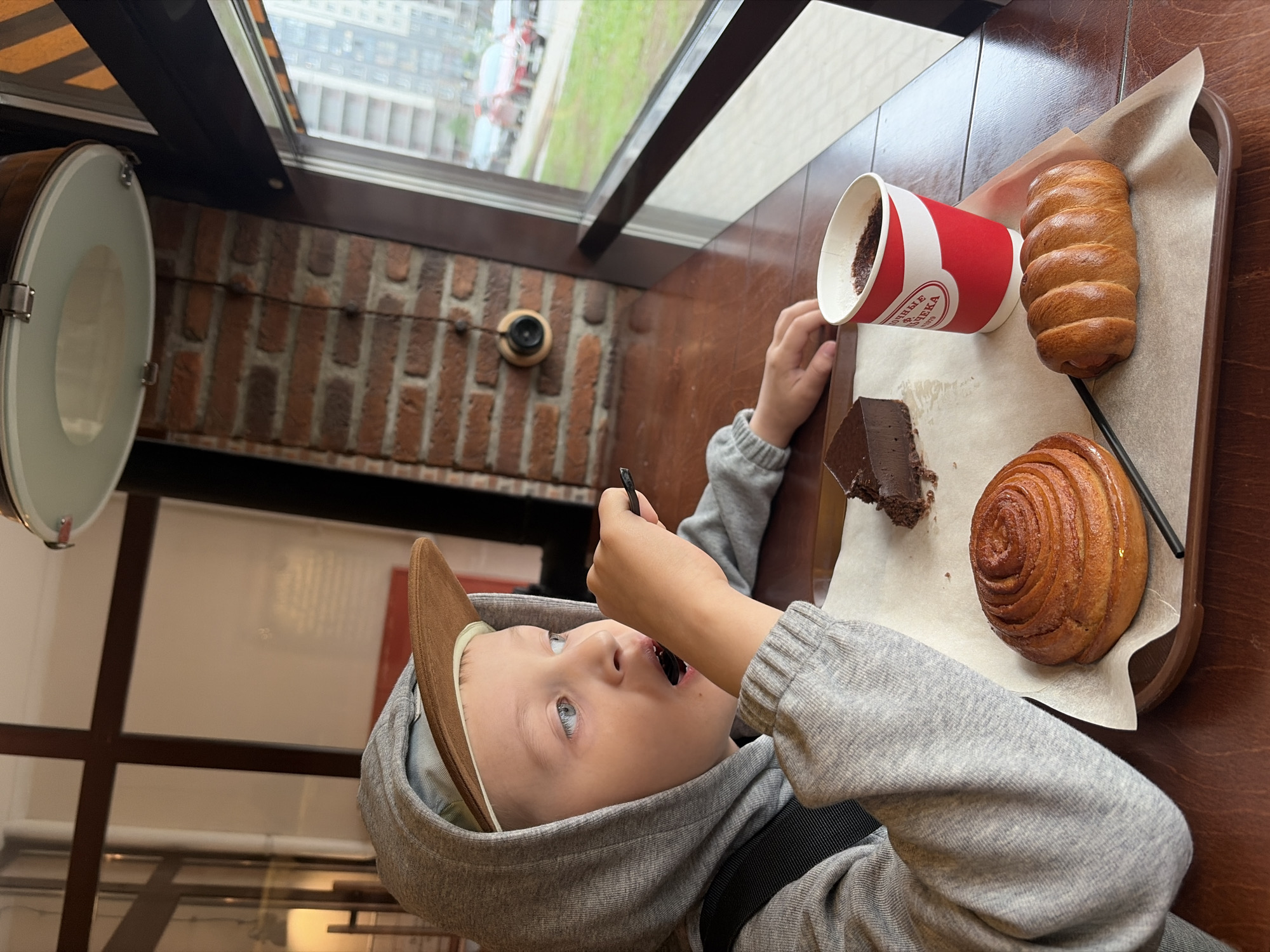 A fair-skinned child with blue eyes, wearing a gray hooded sweatshirt and a brown baseball cap, is eating a piece of chocolate cake with a small spoon. The child sits at a wooden table near a window, with a tray of food in front of them. The tray contains a red and white paper cup of coffee, a chocolate pastry, a cinnamon roll, and a sausage roll. Outside the window is a view of a cityscape with green trees and buildings. The interior setting suggests a cafe or bakery with brick walls and a circular light fixture. The location is Saint Petersburg, Russia, as indicated on the coffee cup.