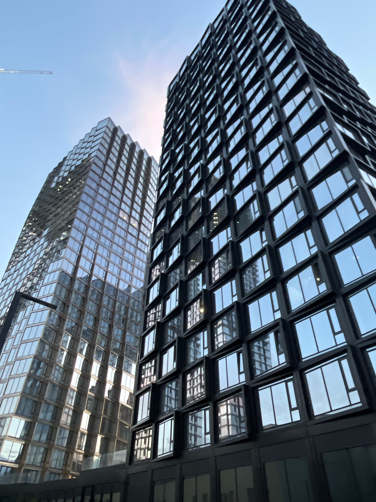 The photograph shows a low-angle view of two modern skyscrapers in Moscow, Russia. The building on the left is clad in glass, reflecting the sky and other surrounding structures, and appears to have a unique, faceted design. The building on the right is darker, with a facade characterized by regularly spaced, dark-framed windows that create a grid-like pattern. A construction crane is visible in the upper left corner, indicating ongoing development in the area. The sky is clear and light blue, with a hint of sun haze visible between the two buildings. The bottom of the image shows the dark facade of a lower level structure or walkway in front of the skyscrapers.