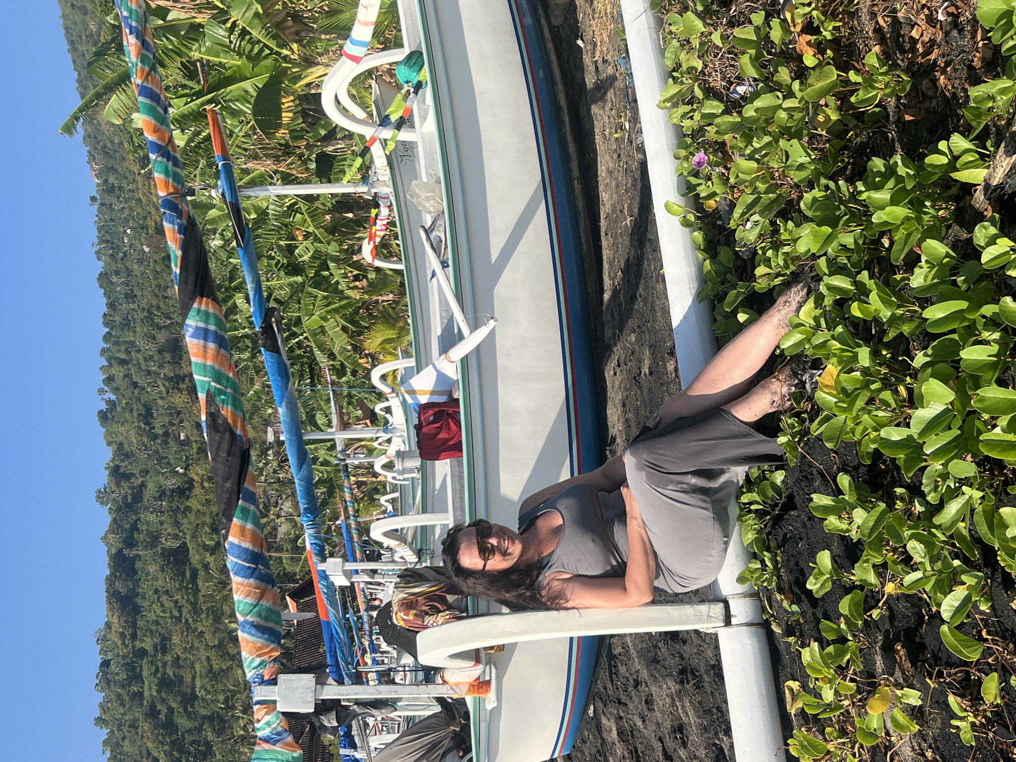 A woman sits on a beach in Abang, Indonesia, next to a row of traditional Indonesian outrigger boats. She is wearing a grey sleeveless dress and sunglasses. The boats are white with blue and red trim, and they are covered with colorful tarps. The beach is made of black sand, and there are green plants growing in the foreground. In the background, a lush green hillside rises steeply, covered with trees and vegetation. The sky is clear and blue.
