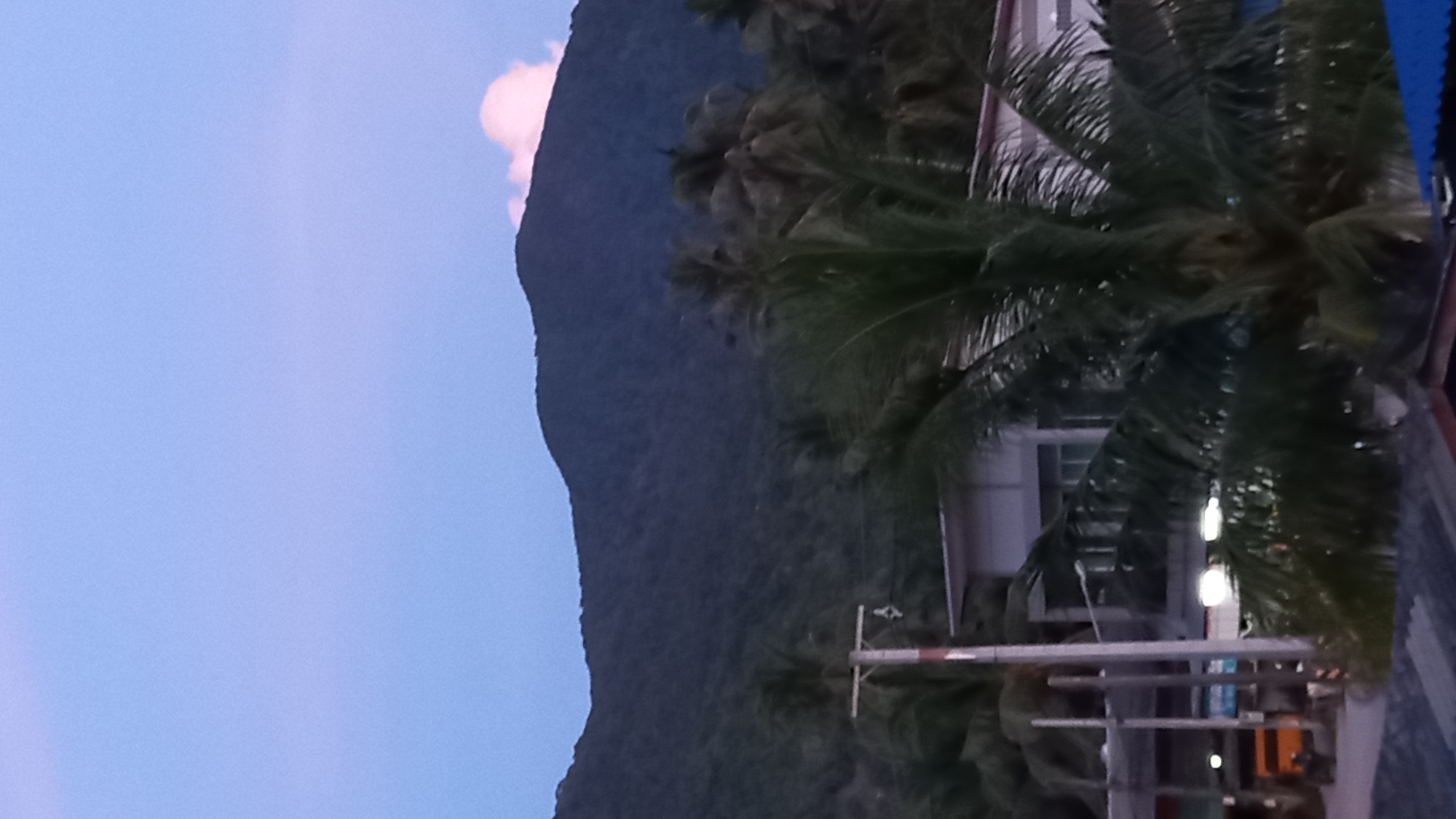 The image shows a view of a town in Tambon Ko Phangan, Thailand, with a mountain range in the background. In the foreground, there are palm trees, a building with a white roof, and a utility pole. A small orange vehicle is visible in the background. The sky is a gradient of blue and purple, suggesting it is either dawn or dusk.