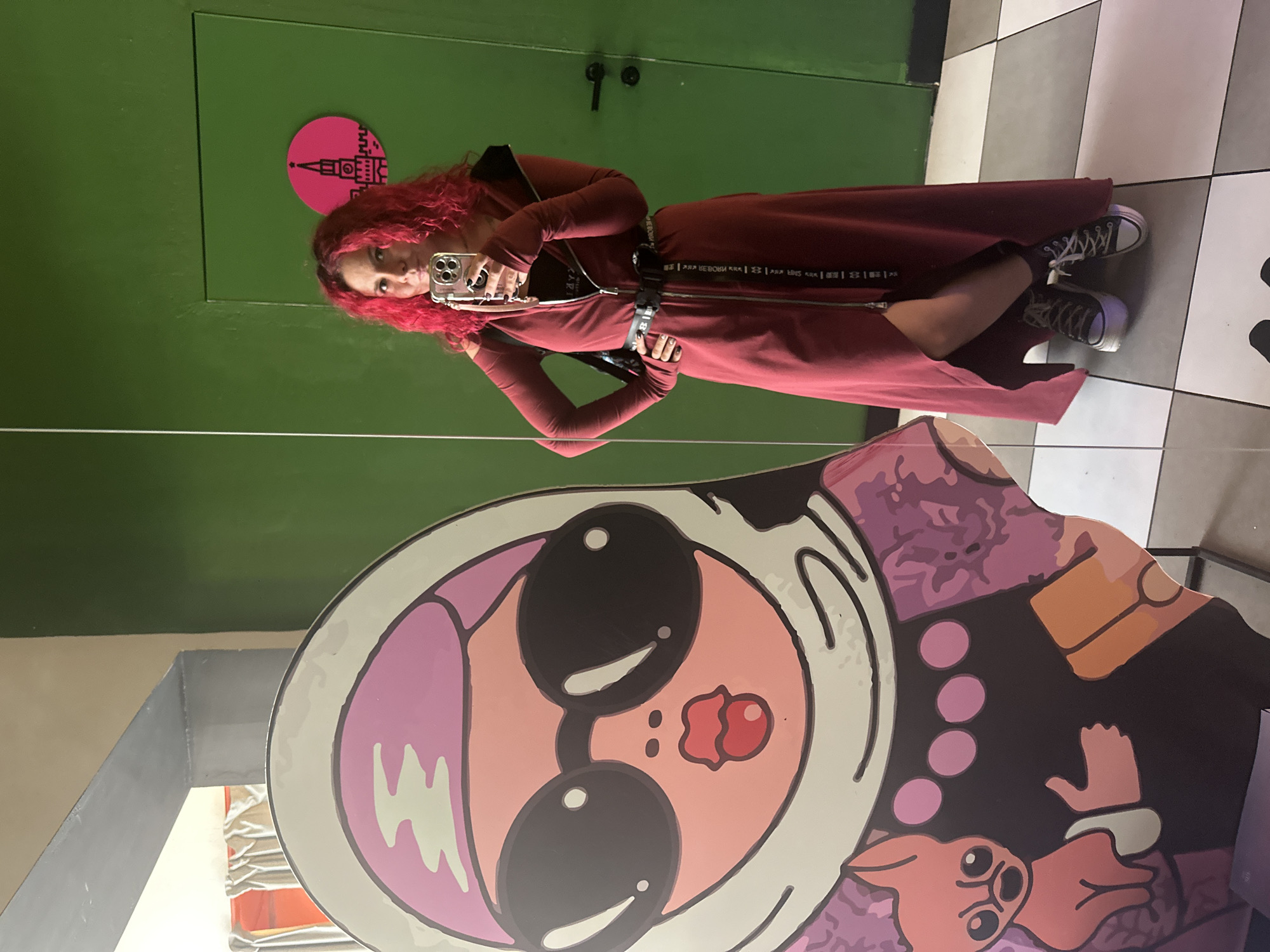 A person with red hair is taking a photo of themselves in a mirror. The person is wearing a long, red zippered dress with sneakers. Behind them is a green wall with a pink circular sign with a black design and a checkered floor. In the foreground, reflected in the mirror, is a large, cartoonish matryoshka doll design. The location is Novosibirsk, Russia, as indicated by the Russian Cyrillic text on the dress.