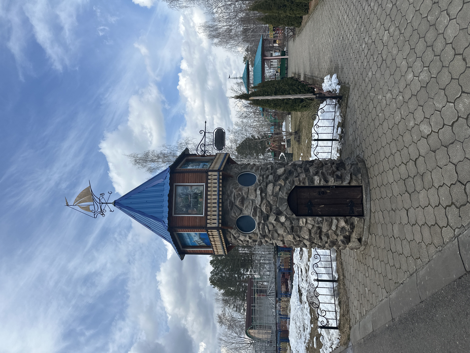This media file depicts a charming, tower-like structure in an outdoor park or recreational setting in Chaikovsky, Russia, under a cloudy but bright daytime sky. The structure has a rustic aesthetic, featuring a stone base with a dark wooden door and two small, round windows. Above the stone, there is a section made of carved wood, housing rectangular windows. One window clearly displays "ЧАСЫ РАБОТЫ" (Opening Hours) and appears to contain a clock face or a painted scene with figures, suggesting a playful or storybook theme. The tower is capped with a vibrant blue conical roof, adorned with a decorative metal weather vane shaped like a sailing ship.
To the left of the tower's upper section, a black wrought-iron sign hangs, prominently bearing the text "УГОЛОК СКАЗОК" (Fairy Tale Corner), reinforcing the whimsical nature of the location. The tower is situated beside a neatly paved pathway made of interlocking grey cobblestones. A low, decorative black metal fence runs along the pathway, bordering areas with patches of melting snow and some sparse vegetation, indicating a late winter or early spring season. In the background, a mix of bare trees and evergreen conifers can be seen, along with another building featuring a teal-colored roof. No people are visible, lending a quiet and serene atmosphere to the scene.