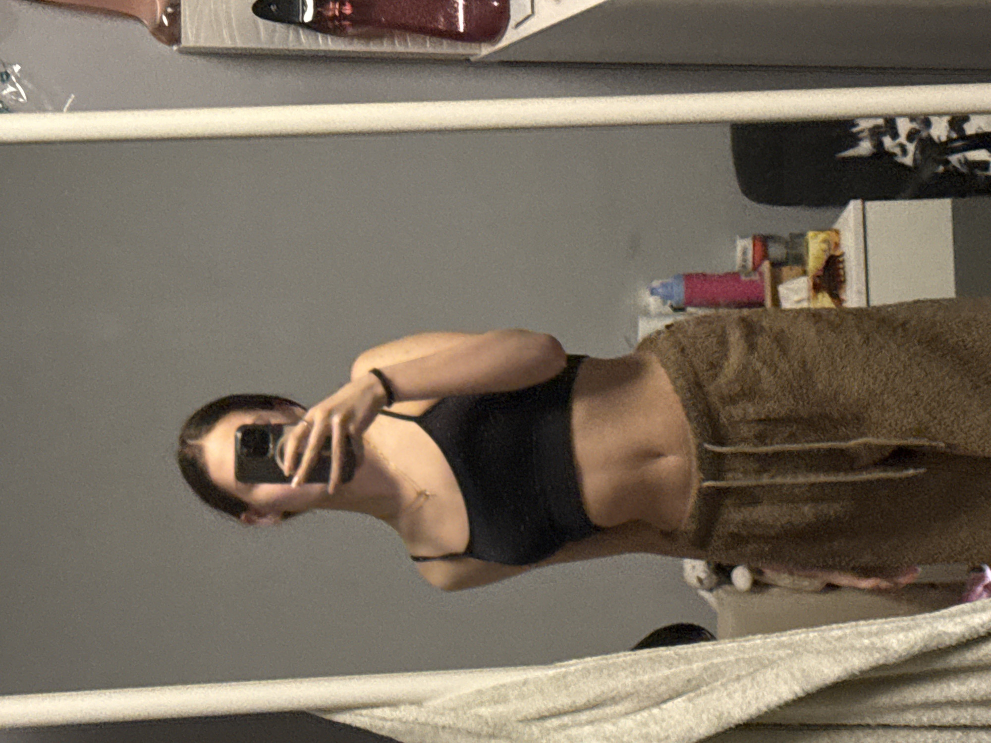 A woman is taking a selfie in a mirror. She is wearing a dark-colored sports bra and brown sweatpants. The background includes a gray wall and a cluttered shelf with various items. The location is not discernible from the image.