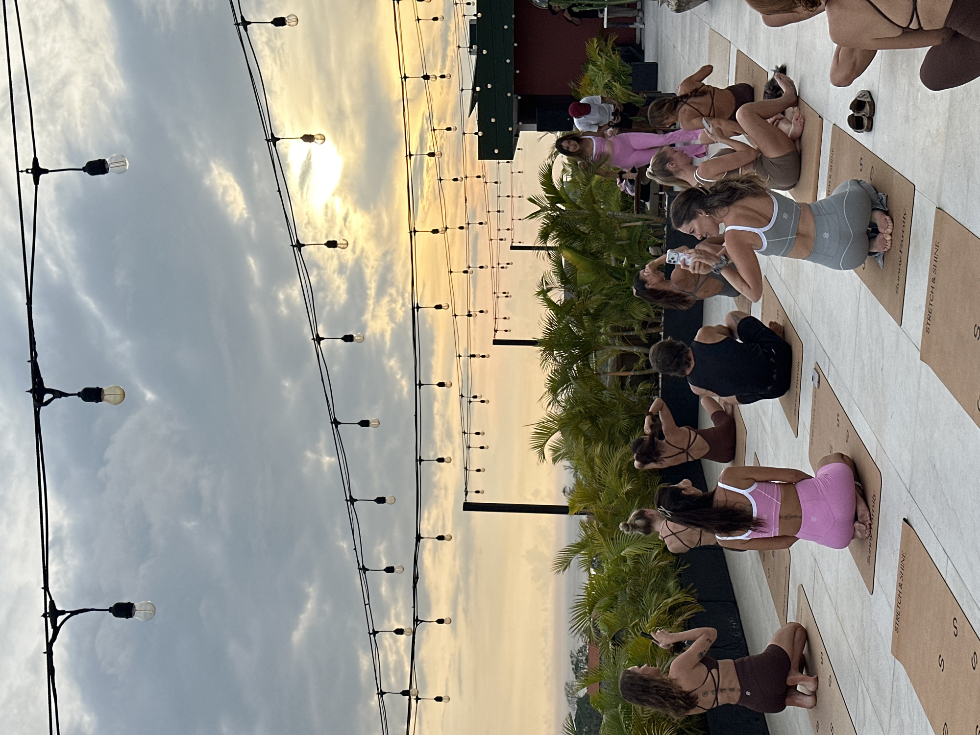 The image depicts a yoga class in progress at sunset in Tibubeneng, Indonesia. A group of approximately ten people are positioned on yoga mats on a paved outdoor surface, likely a rooftop or patio. They are arranged in rows, some holding their phones and looking at them. Palm trees and other tropical foliage are visible in the background, indicating a tropical setting. Strings of lights extend across the top of the scene, suggesting an outdoor event space. The sky shows a mix of clouds and the setting sun. The time of day appears to be late afternoon or early evening, based on the lighting. The yoga mats have a brand name on them.