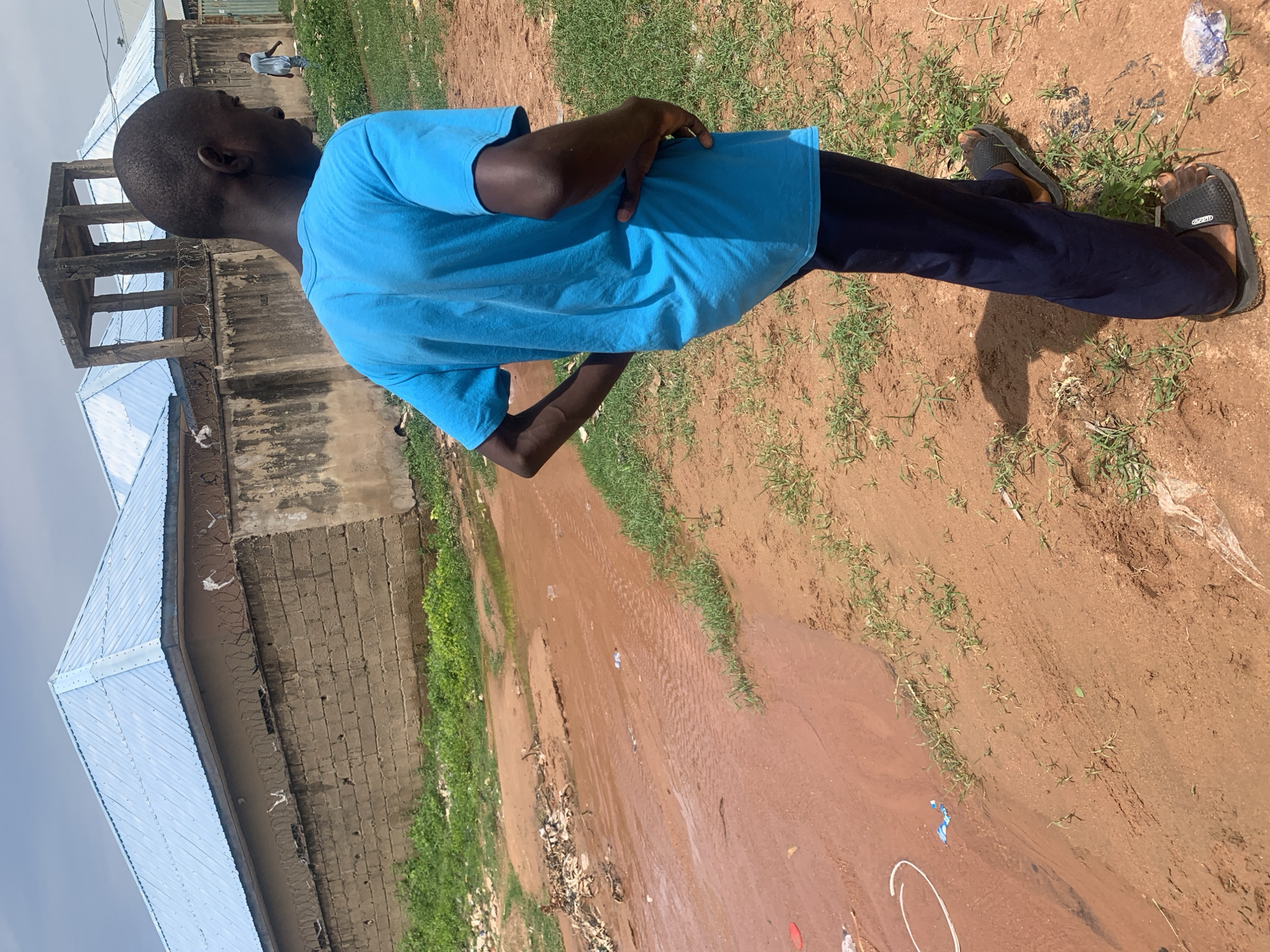An African male wearing a bright blue t-shirt and dark blue pants stands with his back to the camera, looking towards the right. He has his hands on his hips. He is standing on a dirt path next to a muddy stream. In the background, there are two buildings with blue roofs and a concrete structure. The sky is overcast. The scene appears to be in a residential area in Jauro Yinu, Nigeria.