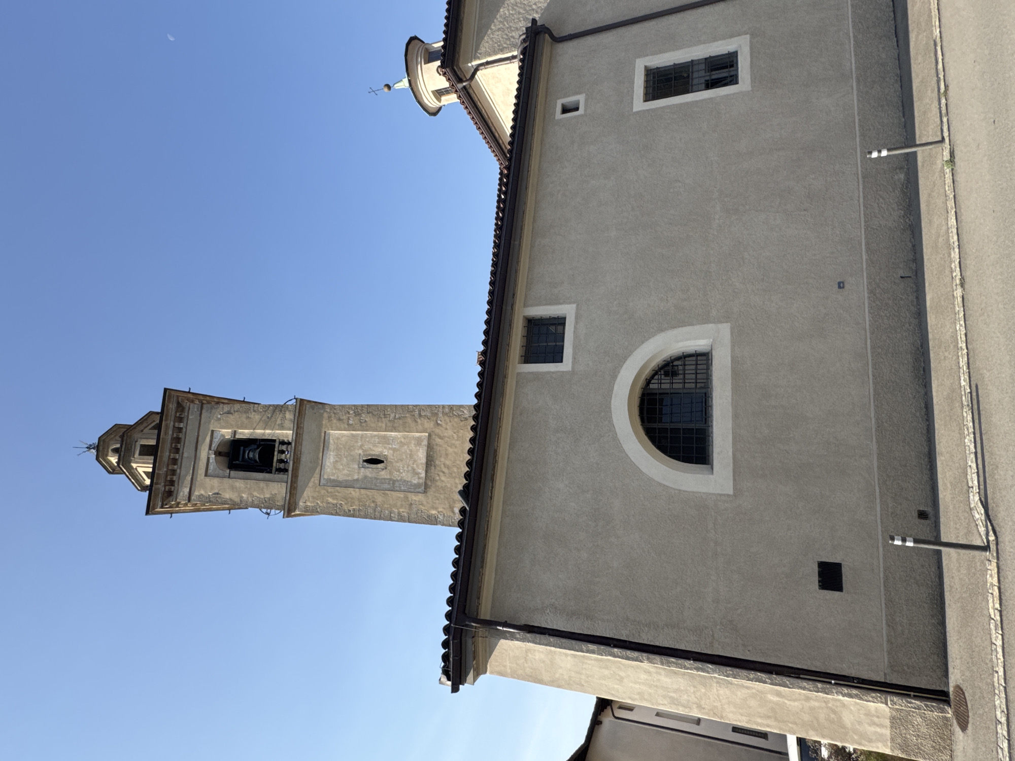 The image shows the Parish Church of San Giorgio in Balerna, Switzerland. The church features a tall, square bell tower with a bell visible inside and a smaller domed structure with a cross on top. The building's facade is a light beige color, with arched and rectangular windows. The surrounding area appears to be a street with a sidewalk and a few bollards. The sky is clear and blue, with a faint crescent moon visible on the right side of the frame. The overall scene is one of architectural significance within a tranquil setting.