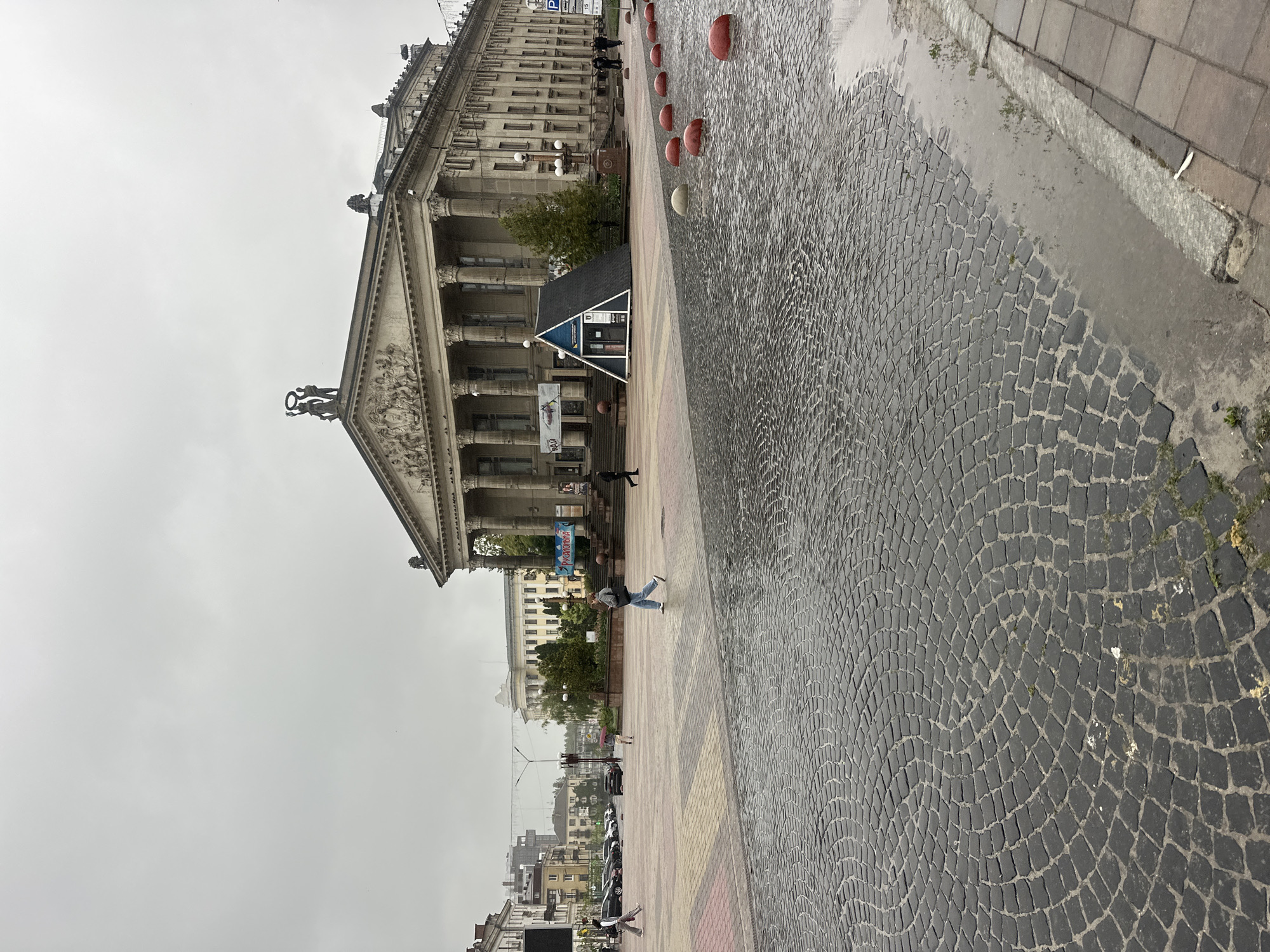 The image captures a wide street or public square in Ternopil, Ukraine, on an overcast day, indicated by the uniformly grey sky and wet ground with visible puddles.
The main subject is a large, imposing building on the left, characterized by its classical architecture. It features numerous tall columns supporting a prominent triangular pediment, which is adorned with intricate relief sculptures. Statues are visible on the roofline and on the pediment's apex, including a sculpture of two figures holding a wreath. This grand structure suggests a significant public, cultural, or administrative institution.
In front of the building, a paved area extends into the foreground, consisting of dark, irregularly shaped cobblestones laid in a concentric circular pattern, alongside smoother sections of multicolored pavement. A line of red, hemispherical bollards runs along the edge of the cobblestone area. Several pedestrians are seen walking across this paved space, going about their day. One person in a light jacket and jeans is captured mid-stride.
Near the entrance of the main building, a small kiosk with a dark roof and blue siding is visible. Text on its side partially reads "IНФОРМАЦIЙНИЙ ЦЕНТР" (Information Center). A large banner with Ukrainian text, including the word "УКРАЇНСЬКА..." (Ukrainian...), hangs near the building's facade, possibly advertising an event or exhibition. In the background, other city buildings and parked cars can be seen under the muted light. A blue parking sign with a white "P" is also visible. The overall scene depicts a quiet, everyday urban environment after or during a period of rain.