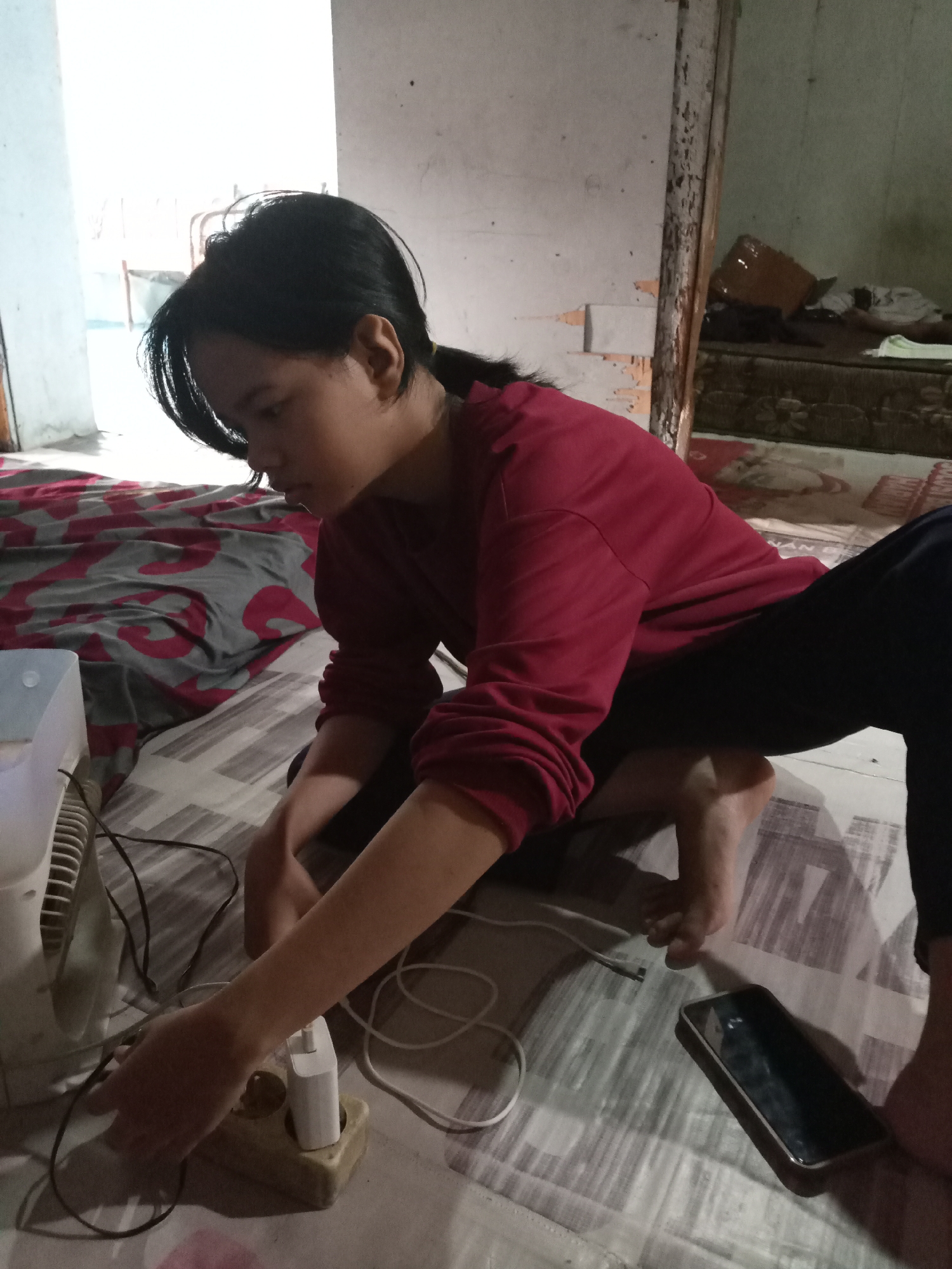 A young Asian female with dark hair pulled back in a ponytail is crouched on a patterned floor, facing away from the camera and towards a white electrical appliance, possibly a fan. She is wearing a maroon long-sleeved shirt and dark pants. Her left hand is reaching towards a tan electrical outlet with two white charging adapters plugged into it, one of which has a white cable connected. A black smartphone lies on the floor to her right. The background consists of a wall with peeling paint and a doorway leading to another room, where a bed with a red and gray patterned blanket is visible. The lighting suggests an indoor environment, potentially in a home in Kapuas, Indonesia.