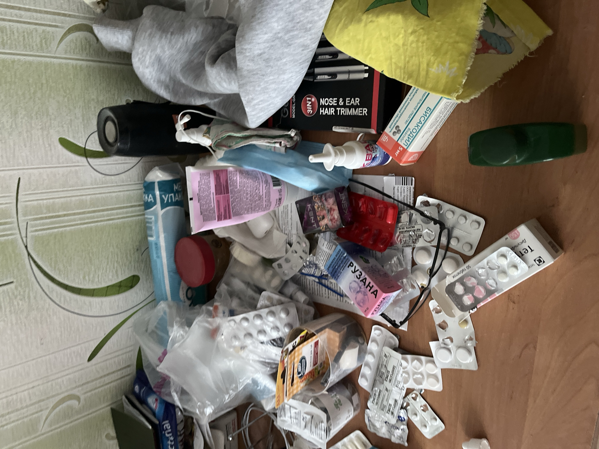 The image depicts a highly cluttered light brown wooden tabletop, situated in what is identified as Minsk, Belarus. The background consists of light green wallpaper adorned with abstract white and green curvilinear patterns. No individuals are present in the scene.
The tabletop is covered with numerous pharmaceutical products and personal effects. Visible medications include multiple blister packs, many of which are partially used or empty, along with boxes for "РУЭАНА" (Rueana), "БИСАКОДИЛ" (Bisacodyl), and "Тен" (Ten, 150 mg tablets). A white nasal spray bottle and a pink tube of cosmetic or dermatological cream are also present.
Additional objects comprise a black cylindrical item, which appears to be a portable speaker or thermos, and a black-framed pair of eyeglasses. White earbuds are observed hanging on the nasal spray bottle. A distinct box for a "3IN1 NOSE & EAR HAIR TRIMMER" is also on the surface. Personal items include a grey hooded garment, a yellow patterned cloth (possibly a towel) with a design of shoes, a red plastic container, and a red-lidded jar. A clear plastic cup contains various small implements. The scene further includes clear plastic bags, various papers and documents featuring Cyrillic text, a blue and white package labeled "BACTERIAL" (possibly wipes or disinfectant), and several cigarette packs with "KENT" branding visible within a red plastic tray.