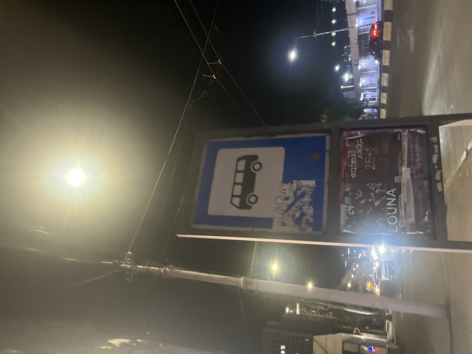 The image shows a bus stop in Yerevan, Armenia, at night. The bus stop sign features a blue and white rectangle with a bus icon. Adjacent to the sign are posters with text and imagery. The background includes streetlights, buildings, and a street with vehicles. Electrical wires are visible in the sky.