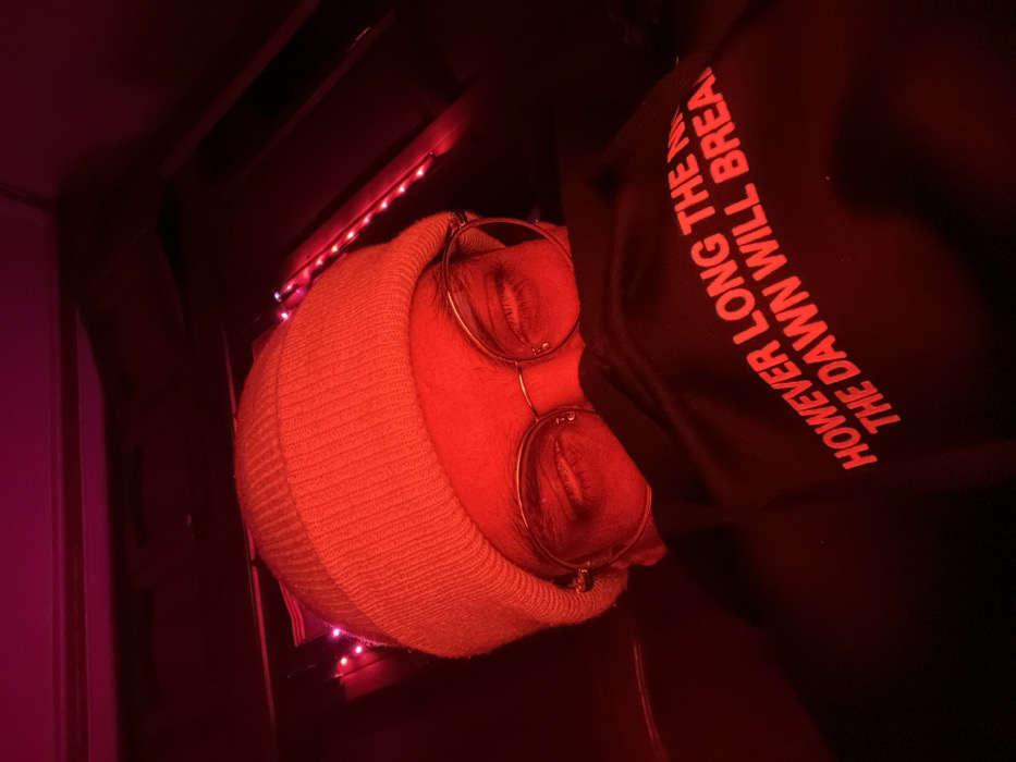 A person with closed eyes wearing a beige beanie and round glasses is illuminated by red light. They are wearing a dark sweatshirt with the words "HOWEVER LONG THE DAWN WILL BREAK" printed in red. Red LED lights are visible along the edge of a shelf or surface. The image is rotated counterclockwise and dominated by red hues, giving it a warm, intense feel.