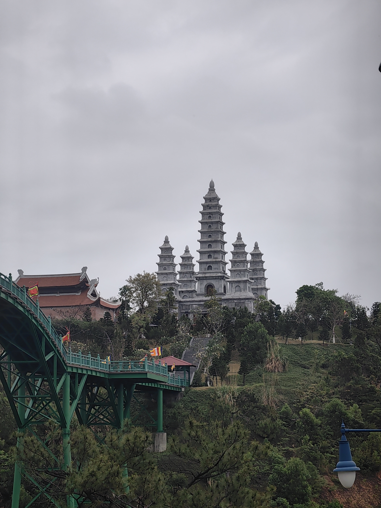 The image captures a view in Hong Gai Ward, Vietnam, featuring a multi-tiered gray stone pagoda complex situated on a hill. The pagoda stands prominently against an overcast sky. Lush greenery surrounds the complex. A green, arched bridge stretches into the foreground, with small Vietnamese flags visible along its railings. A section of a building with a red tiled roof can be seen partially obscured by trees near the pagoda complex. In the lower right corner, a blue streetlamp is present. The overall scene appears tranquil.