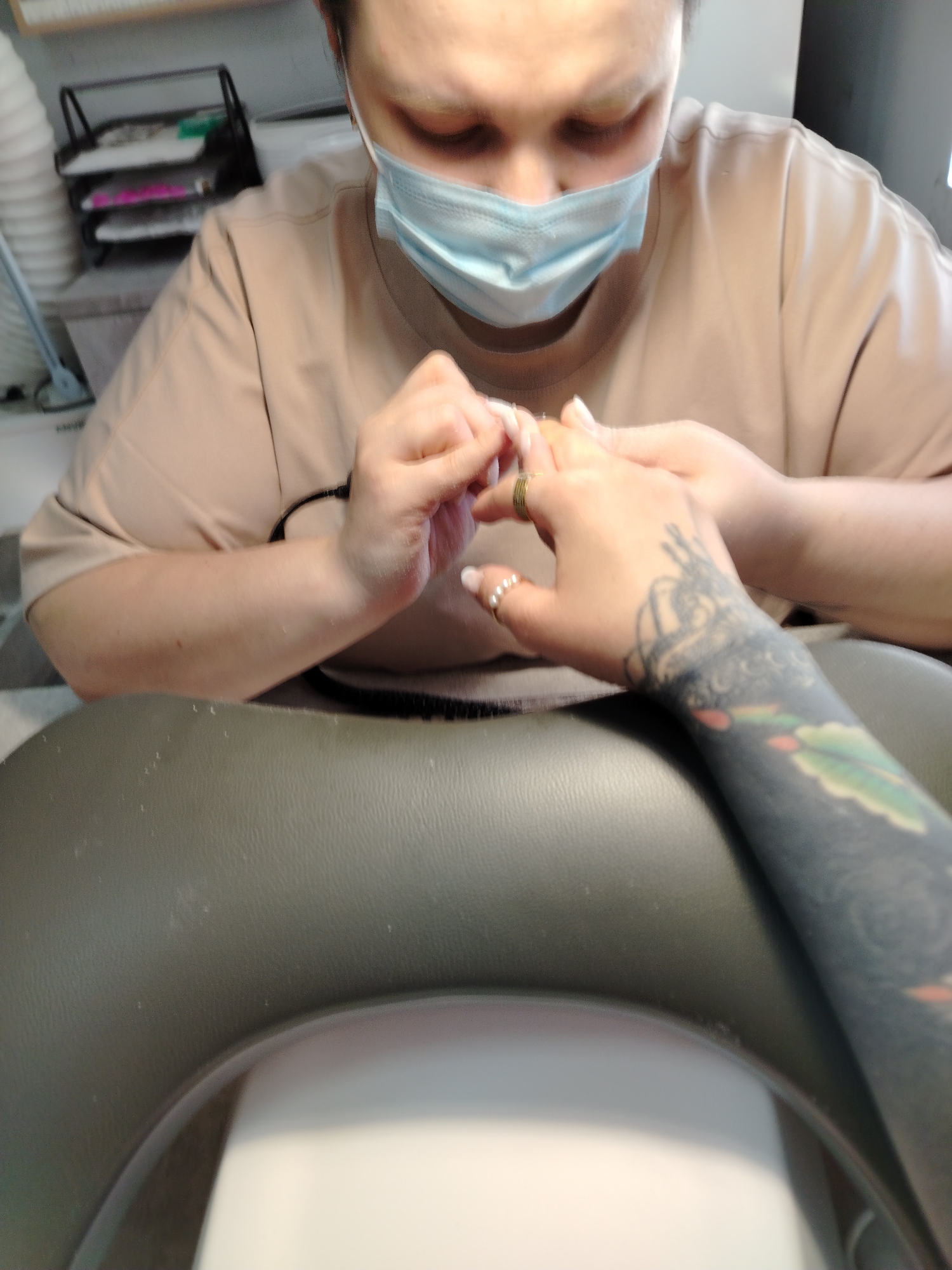 A person wearing a face mask and a light brown shirt is working on someone's nails. The client has a tattoo on their arm and is wearing rings on their fingers. The scene appears to be taking place in a nail salon. There is a grey, padded surface below the client's arm, possibly a manicure table. A shelf with various items on it can be seen in the background. The location in the city of Noviy, Russia, cannot be determined from the visual information provided.