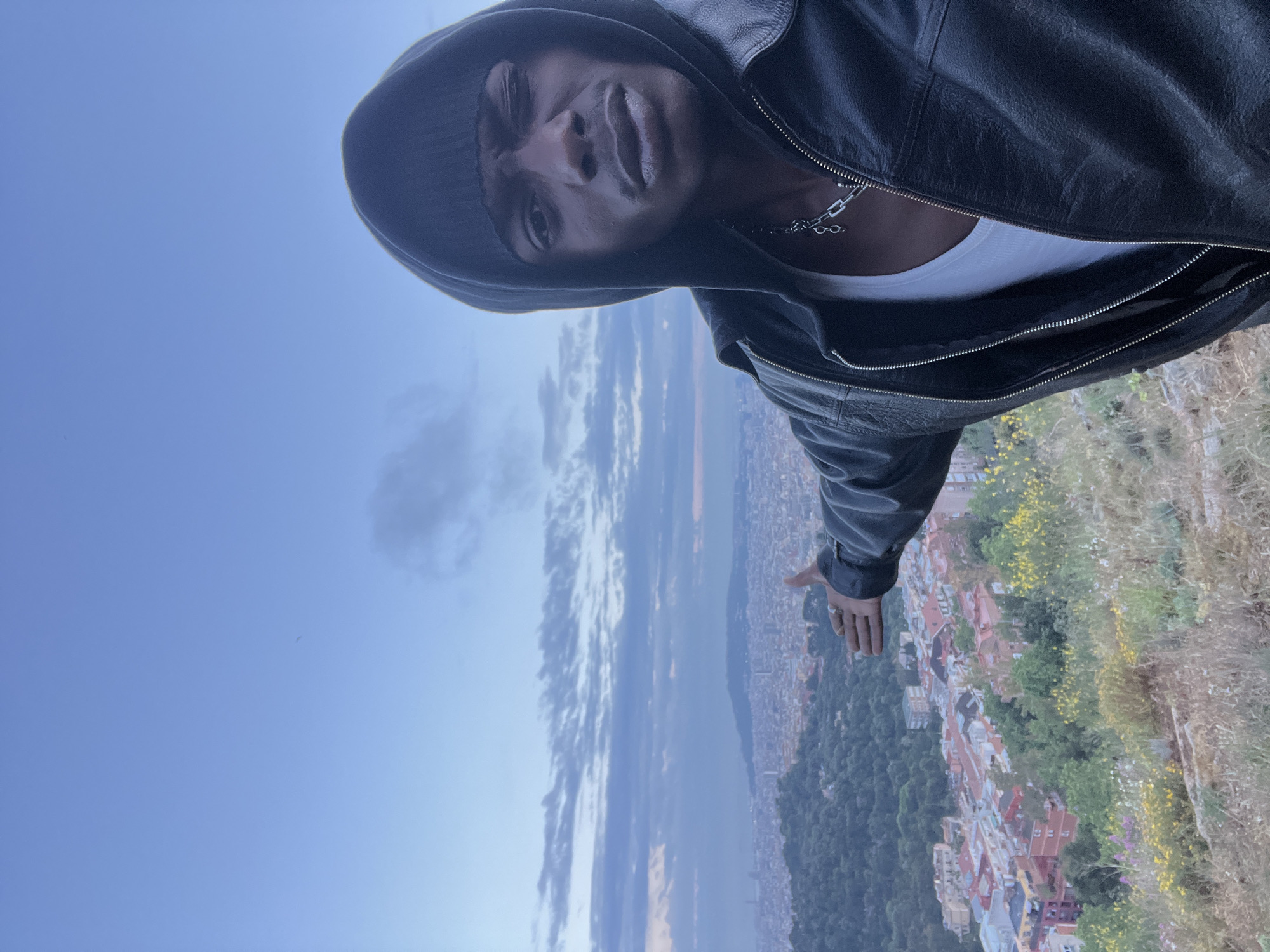The image depicts a person standing atop a hill overlooking the city of Barcelona, Spain. The individual is facing the camera and wearing a black hooded jacket and a knit cap. A silver chain necklace is visible against a white undershirt. The city of Barcelona is in the background, partially obscured by trees on the hillside. The sky is a gradient of blue. The image appears to have been rotated counterclockwise.