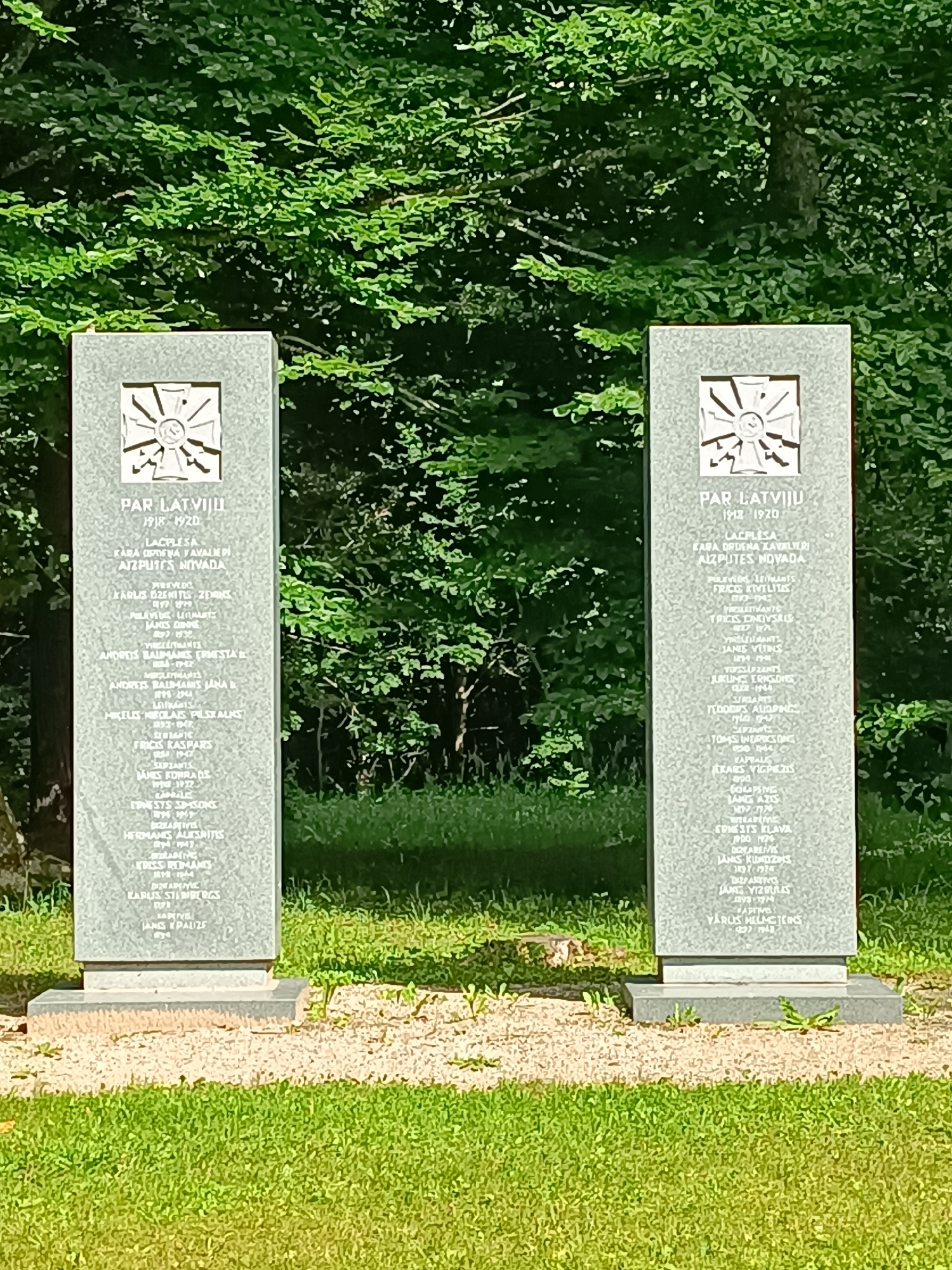 Two granite monument pillars stand side-by-side in a grassy area with a dense green forest backdrop. Each pillar is rectangular and oriented vertically, with a carved emblem at the top and lists of names inscribed below. The left pillar is slightly closer to the viewer. The pillars are likely part of a memorial. The inscription "PAR LATVIJU 1918-1920" is visible on both. The ground in front of the pillars is covered with gravel and grass. Sunlight illuminates the scene, creating shadows on the grass. The overall impression is one of solemn remembrance within a natural setting, possibly a cemetery or a public park.