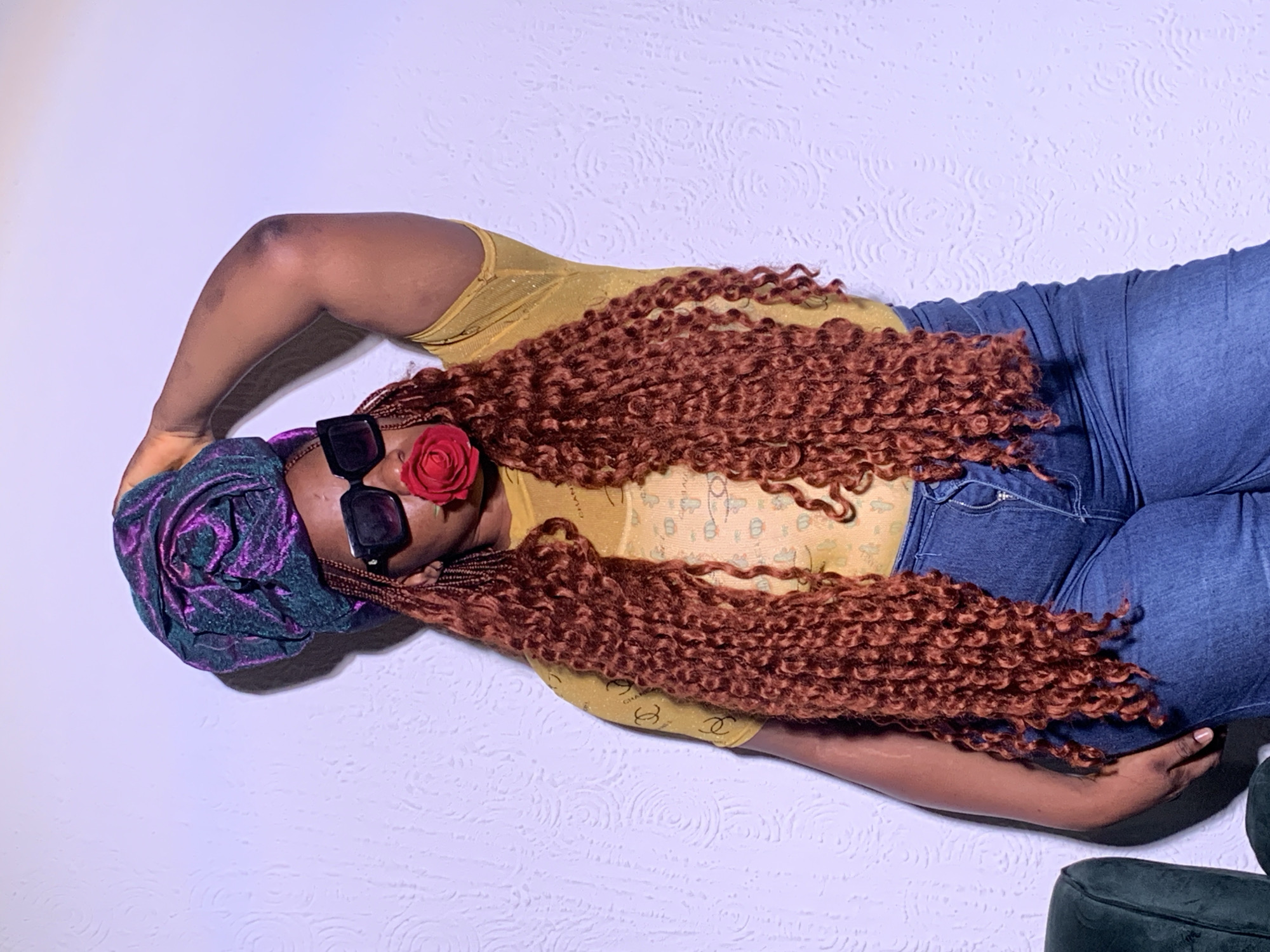 A woman is depicted lying down against a light-colored, textured wall in an indoor setting in Barakin Akawo, Nigeria. She is dressed in a yellow top and blue denim jeans, with a dark purple and green patterned headwrap covering her head. Her long, reddish-brown curly hair cascades over her shoulder and chest. She wears large, dark, square-framed sunglasses, and a red rose is tucked behind the left lens. One arm is bent above her head, and her legs are also bent. The yellow top features a faint, repeating pattern of the word "CHANEL". A dark, upholstered piece of furniture is partially visible in the bottom right corner of the frame. The lighting indicates an indoor environment, and her facial expression is obscured by the sunglasses and rose, suggesting a relaxed or contemplative pose.