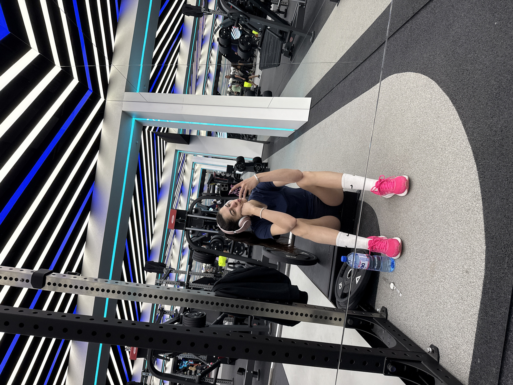 A woman is sitting on the floor in a gym, taking a selfie. She is wearing a navy blue t-shirt, black shorts, white socks, and pink sneakers. She has headphones on. Next to her is a water bottle and a black jacket. The gym has a mirror on the wall, reflecting the woman and the gym equipment behind her. The walls and ceiling have a pattern of black, white, and blue stripes. The floor is gray and black. Gym equipment is in the background, including a squat rack and leg press. The specific location is not discernible from this image.