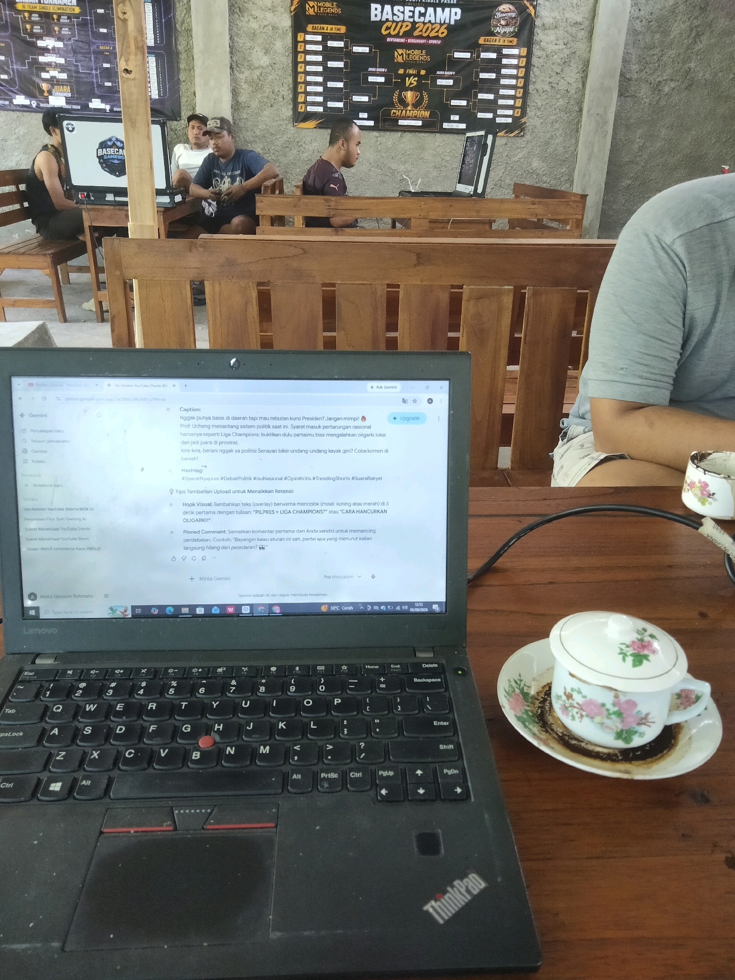 A medium shot captures a laptop open on a wooden table, displaying a webpage with text and images. To the right of the laptop, a teacup and saucer with a floral pattern are placed. In the background, several individuals are seated at wooden tables and chairs, appearing to be in a cafe or an event venue. Two tournament posters are visible on the wall behind them, one featuring "BASECAMP CUP 2026" and another with "MOBILE LEGENDS." The environment suggests an esports or gaming-related gathering. The presence of the tournament posters and people watching screens implies the location is likely a gaming center or a place hosting a gaming tournament in Merakurak, Indonesia. The overall atmosphere is casual and focused on the event.