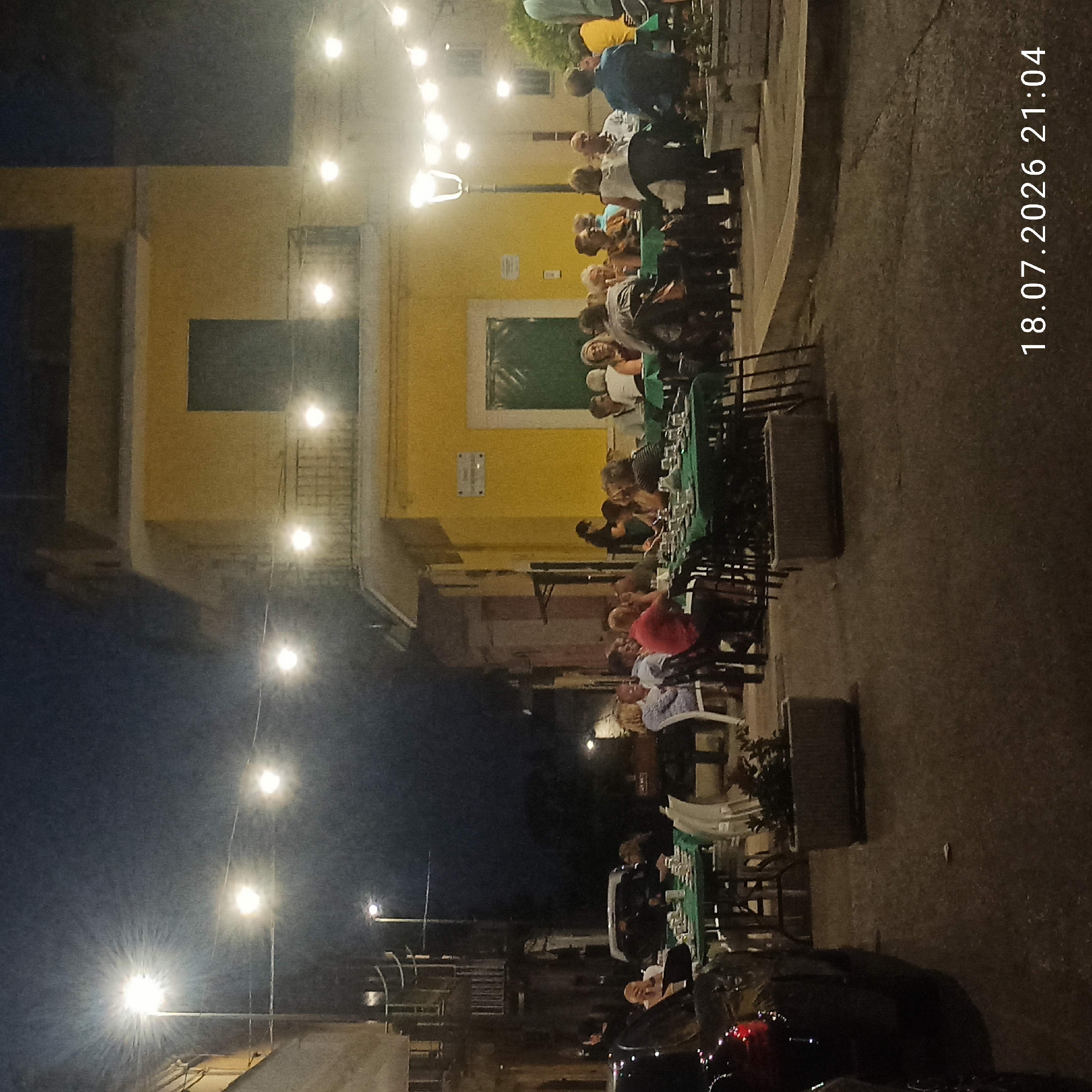 The image captures an outdoor dining scene at night in Bitonto, Italy. The setting appears to be a street or piazza, with a yellow building dominating the background, featuring a green-shuttered window and a balcony. String lights and a street lamp illuminate the area, casting a warm glow on the tables set up for patrons. Several groups of people are seated at tables covered with green tablecloths, engaged in conversation and dining. The patrons are diverse in age and gender, dressed in casual evening attire. Some are looking towards the camera, while others are interacting amongst themselves. In the foreground, a dark car is parked on the left, and planters with greenery are visible, adding to the ambiance of the outdoor restaurant. The ground is paved with asphalt and appears slightly wet in places. The timestamp "18.07.2026 21:04" is visible in the bottom right corner, indicating the date and time the photo was taken.