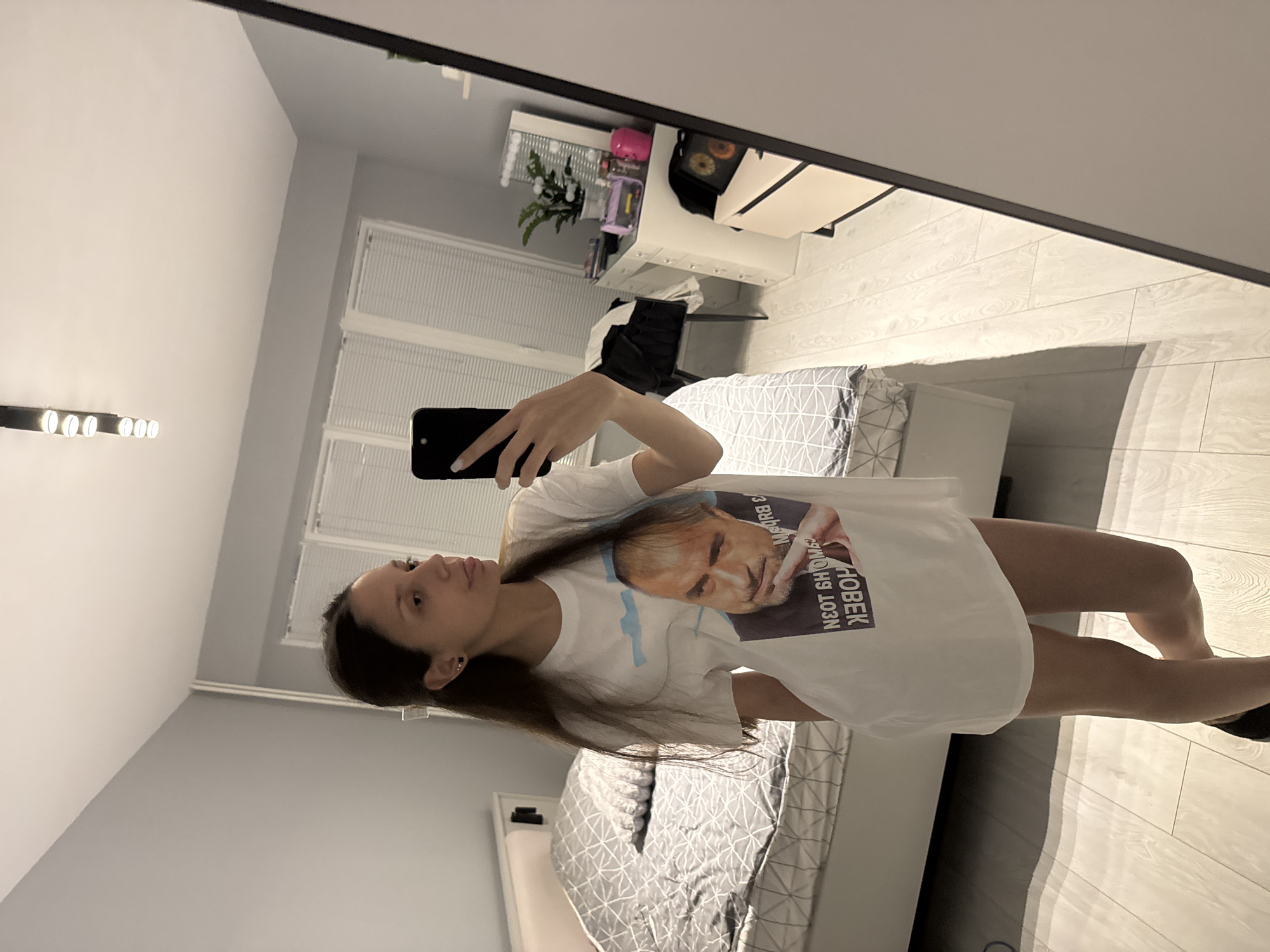 A young woman is taking a mirror selfie in what appears to be a bedroom. She is holding a black smartphone in her left hand and is wearing a white T-shirt with an image of a man on it. The room contains a bed with a grey patterned bedspread, a desk and chair, and what appears to be track lighting on the ceiling. The exact location within Sofia, Bulgaria, cannot be determined from the provided image.
