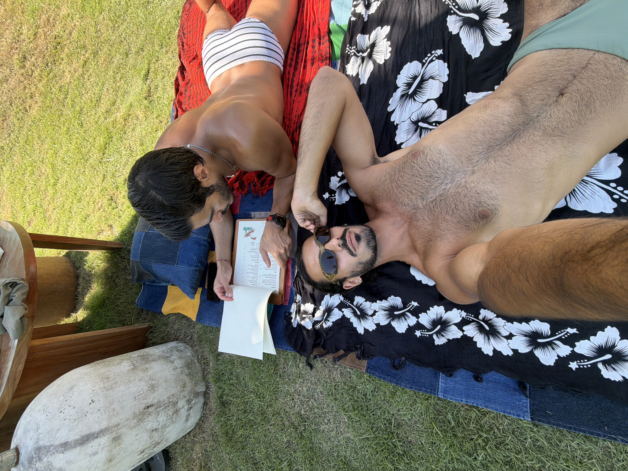 Two shirtless men are lying on a blanket on the grass. One is wearing white and black striped swimwear, and the other is wearing sunglasses and light green swimwear. They are looking at a menu together. A small table with a white, stone-like object next to it is visible. The scene is in Mengwi, Indonesia.