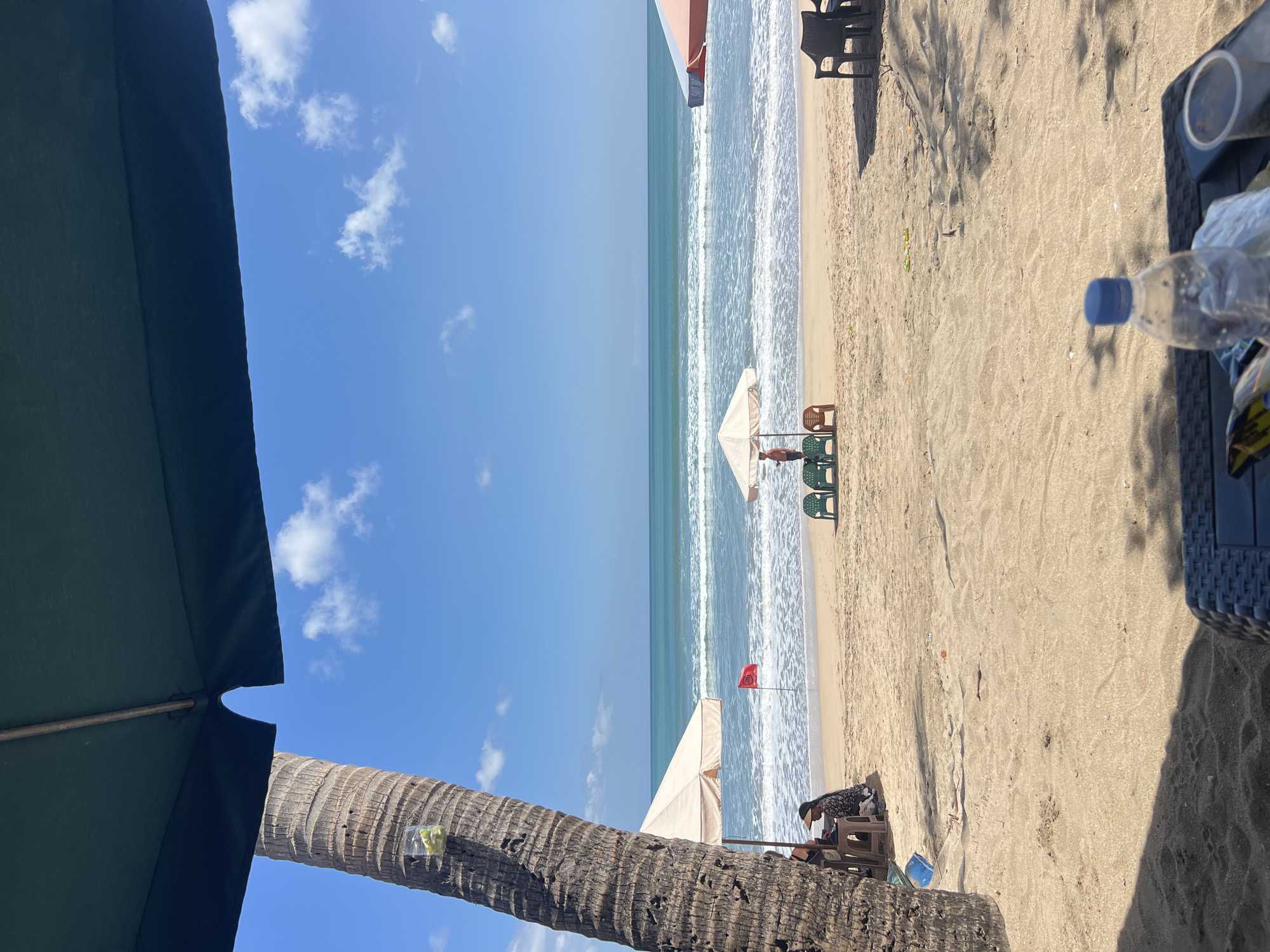 The image captures a serene beach scene in Kuta, Indonesia, on a clear, sunny day. In the foreground, a large dark green umbrella partially obscures the view, with the textured trunk of a palm tree to the left. A glass with a light-colored liquid and some green items is attached to the palm tree. The beach itself is sandy, with gentle waves rolling onto the shore.
Several beach chairs and umbrellas are set up. Two chairs and a small table are positioned under a beige umbrella to the left, where a person wearing a hat is seated, seemingly engaged with something on the table. Further down the beach, a red flag is planted in the sand. In the middle ground, a person stands next to a beige umbrella, surrounded by three green chairs and one brown chair. Another umbrella, partially visible on the right, is orange and white. Dark chairs are also visible on the right side of the frame. In the lower right corner, a table with a bottle of water and some other items suggests a seating area. The ocean stretches to the horizon under a bright blue sky dotted with white clouds. The overall impression is one of a relaxed beach environment, characteristic of Kuta.