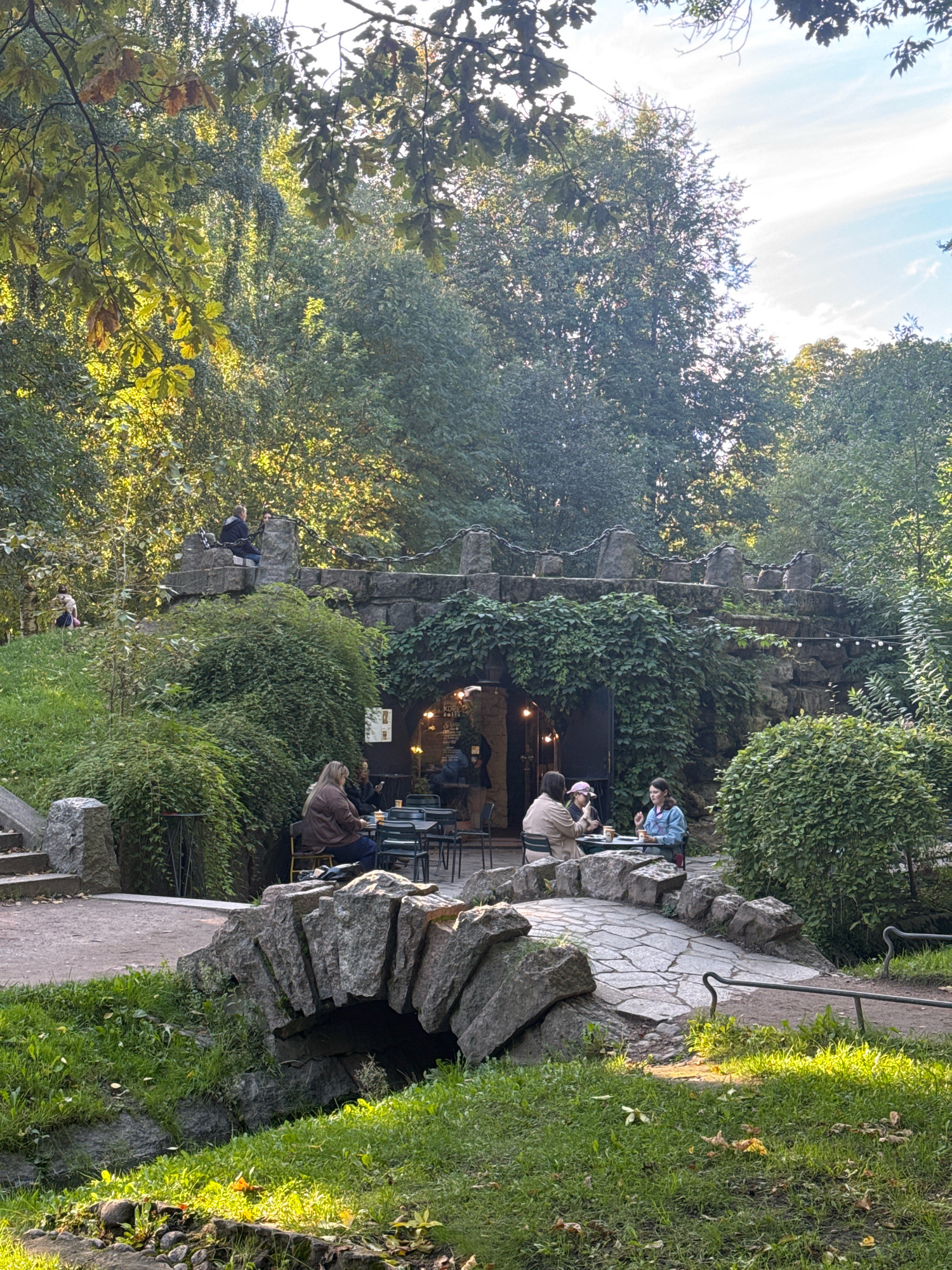The image depicts a scene within a park, likely a public garden or recreational area, with a small stone bridge spanning a narrow channel of water in the foreground. A stone structure, appearing to be a building or a cafe entrance, is nestled into a hillside covered in lush greenery, with stone masonry and an arched entrance. Several individuals are seated at tables outside this structure, suggesting a cafe or dining area. The surrounding environment is characterized by mature trees with dense foliage, indicating a natural or semi-natural setting. The time of day appears to be afternoon, with sunlight casting dappled shadows and highlighting the greenery. The overall impression is one of a serene, outdoor gathering space within a park. It is highly probable that this location is within St. Petersburg, Russia, given the architectural style of the stone structures and the prevalence of such parkland within the city.